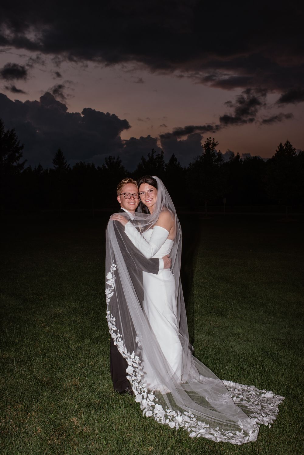 A wedding couple embracing on a grassy field at dusk with a dramatic cloudy sky in the background.