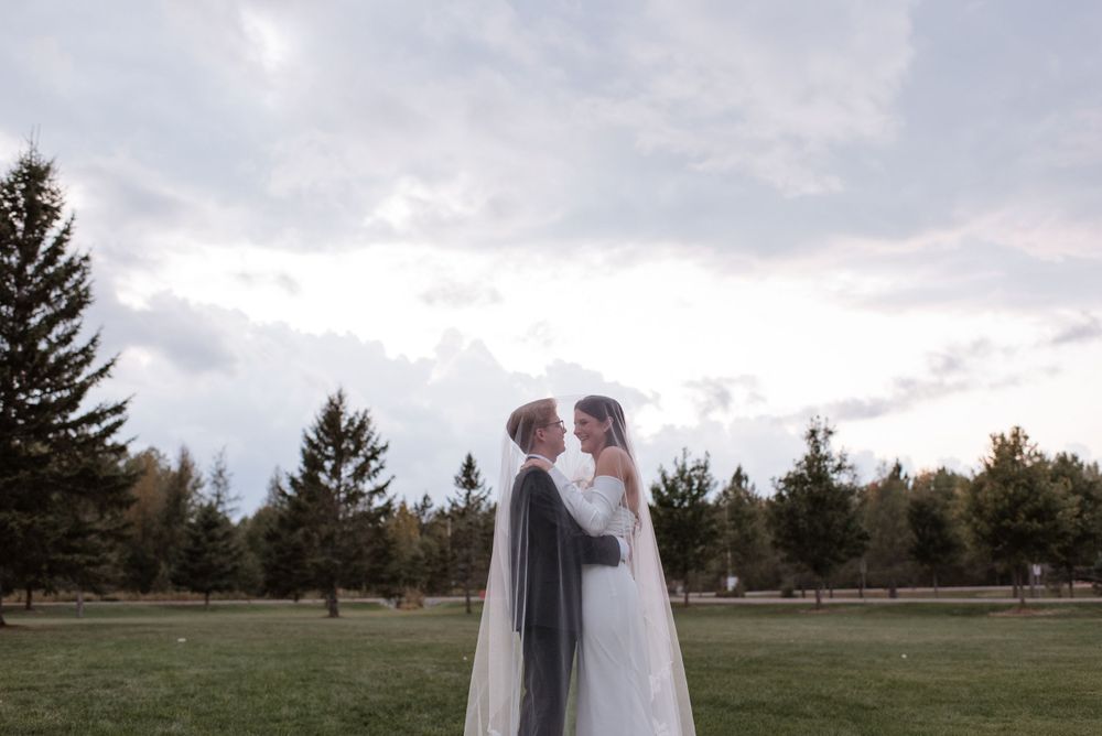 A romantic moment between newlyweds standing in an open field with evergreen trees and cloudy skies in the background.