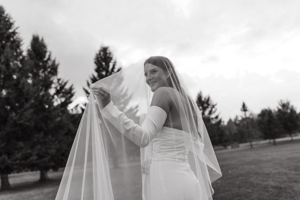 A bride in a white dress adjusts her long flowing veil outdoors with evergreen trees in the background.