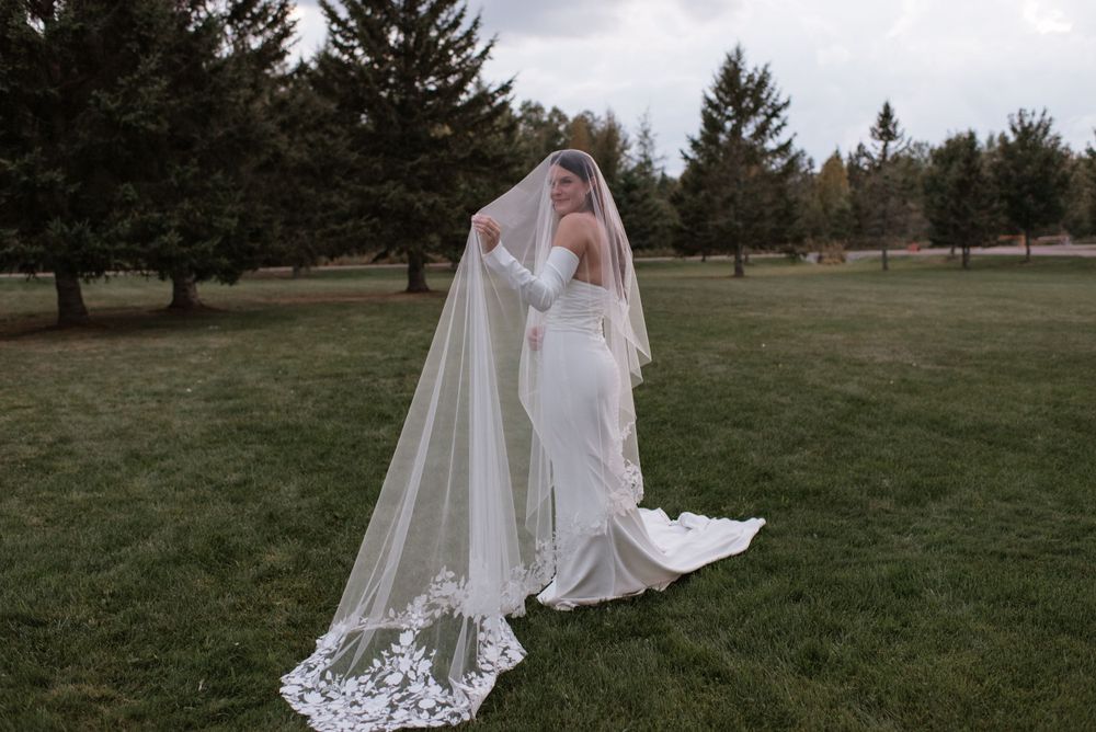 A bride in an elegant white dress with a flowing lace veil stands on a grassy lawn surrounded by evergreen trees.