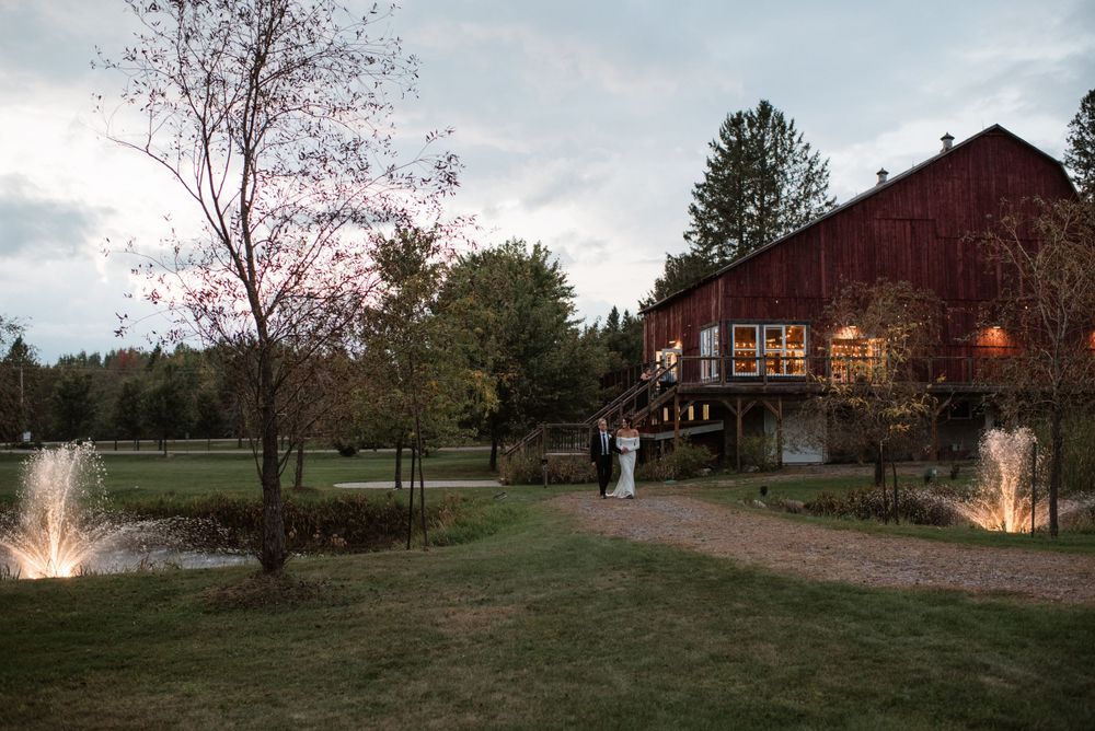 A rustic red barn venue with glowing lights sits near a tranquil pond at dusk with trees surrounding the landscape.