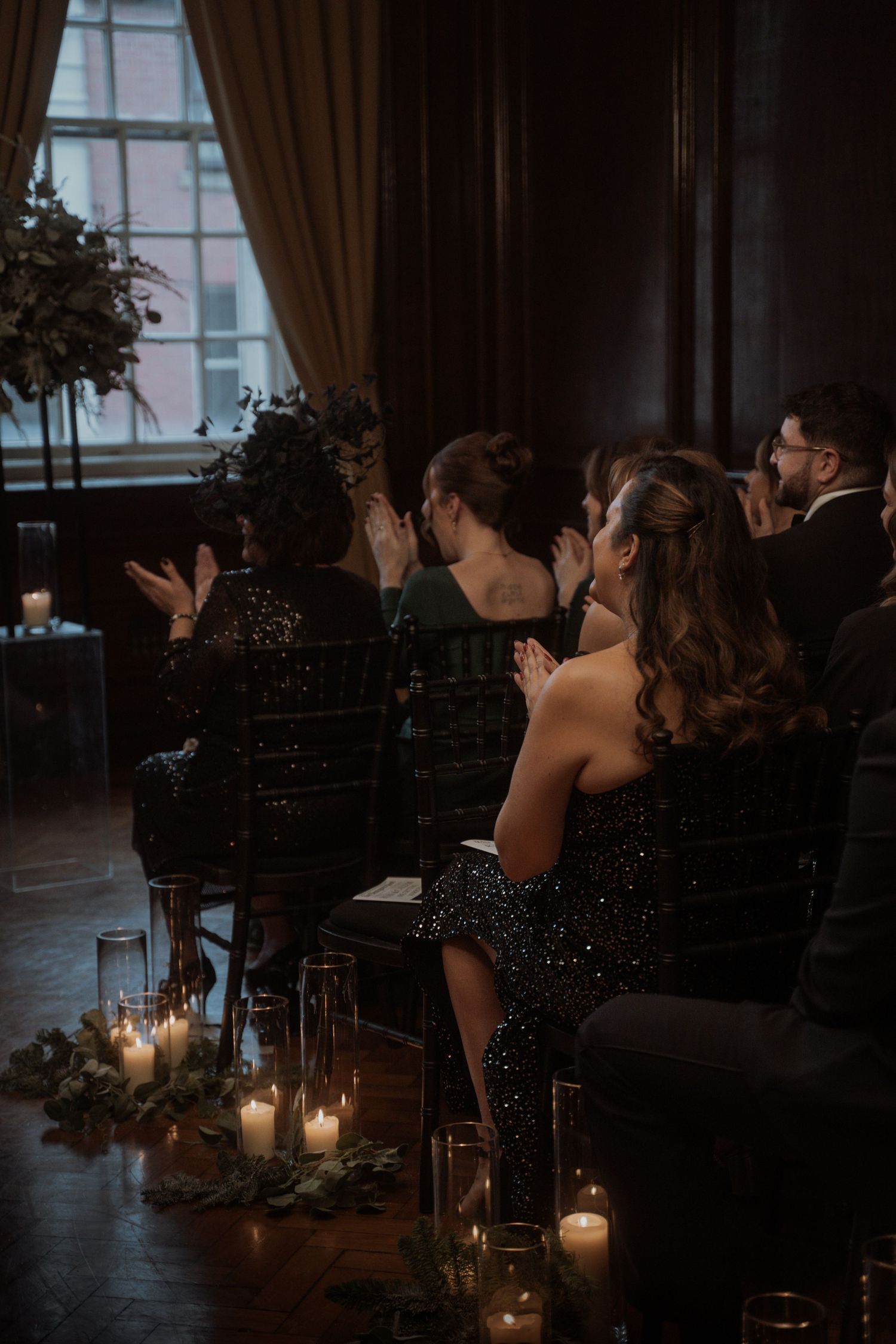 Wedding guests seated in elegant formal attire watch a ceremony by candlelight in a dark, romantic venue.