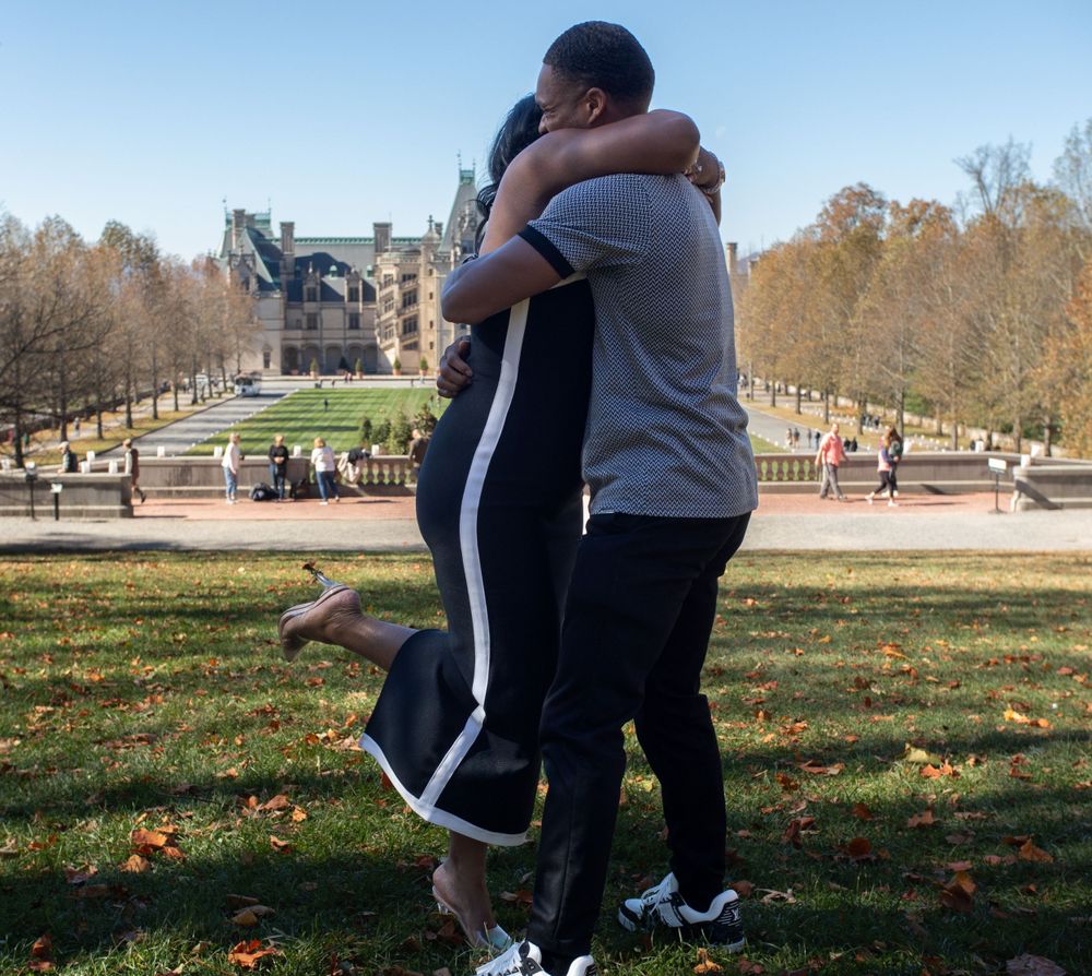 A couple embraces on grass with a grand building visible in the background.