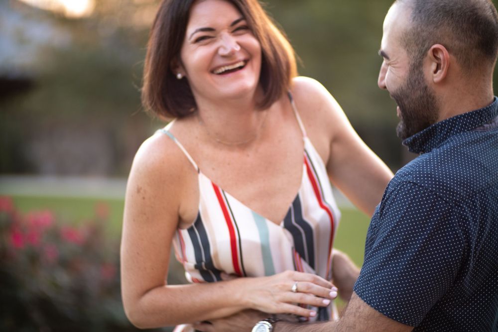 Couple sharing laughs outdoors with woman in striped dress and rose garden in background.