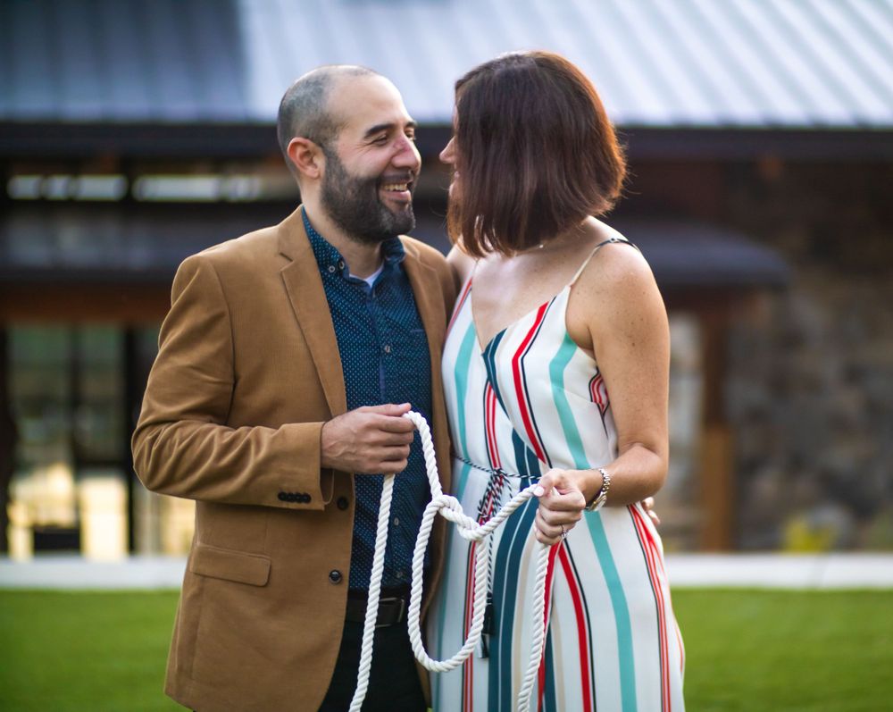A couple shares a joyful moment on a lawn, with the woman wearing a striped dress and man in a tan blazer.