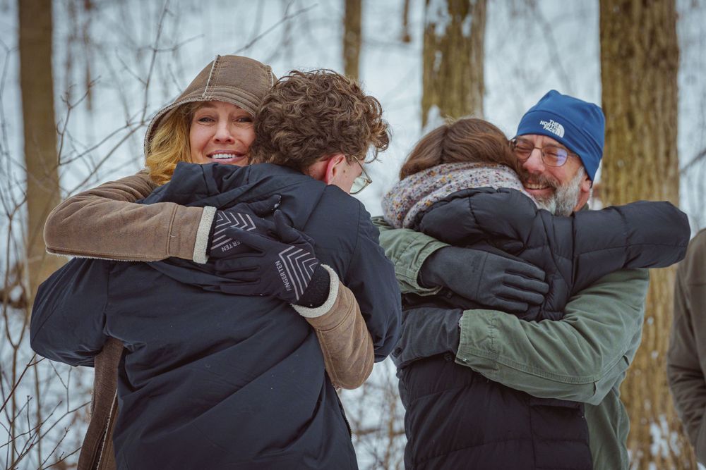 A newly engaged couple share a warm embrace with the fiancée's parents in a snowy winter forest setting after a proposal.