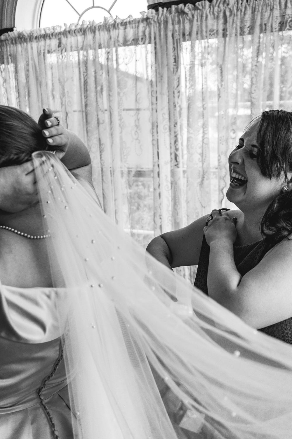Black and white photo of bridesmaids laughing while adjusting a wedding veil.