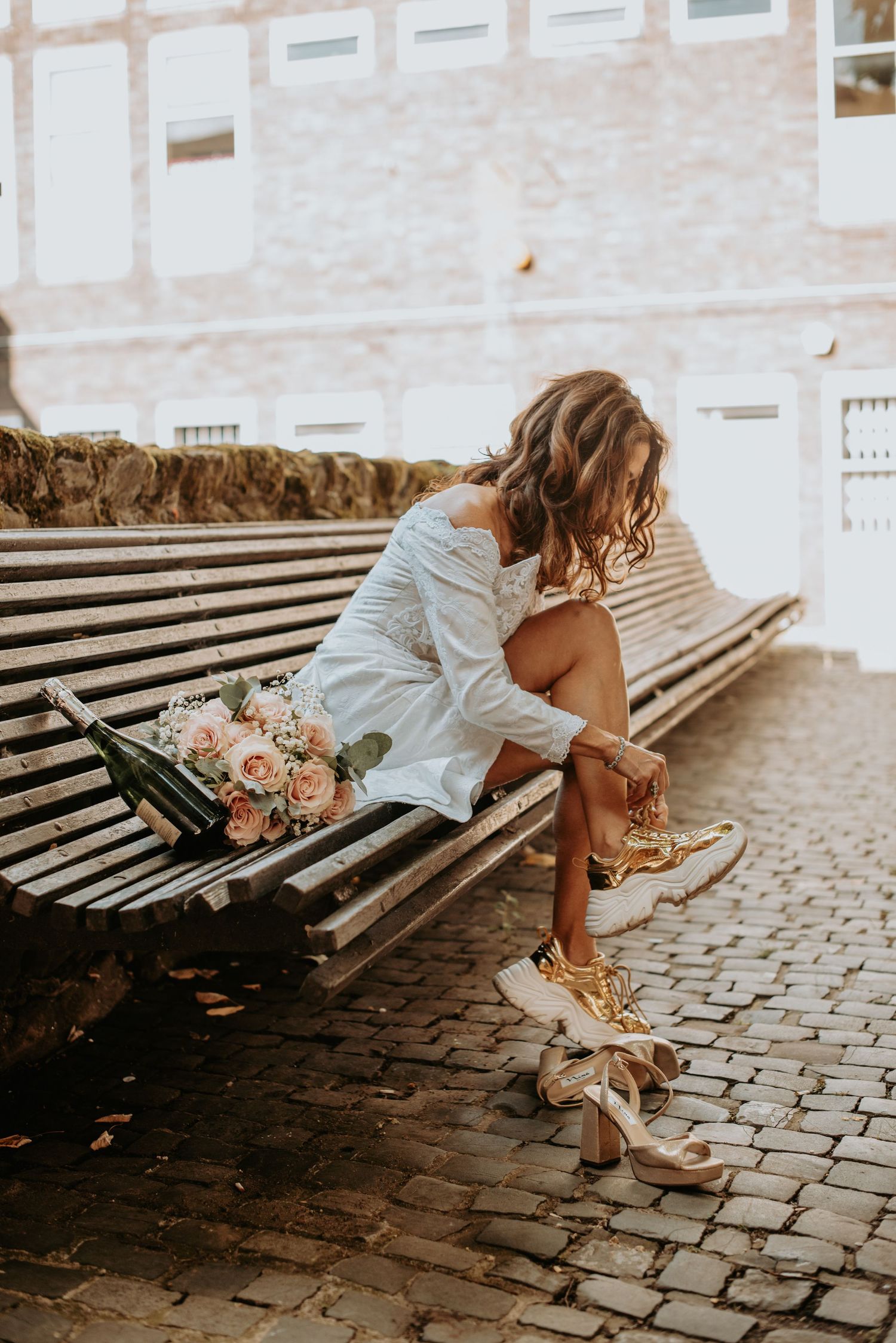 A white-dressed figure sits on a wooden bench with a pink bouquet beside them on a cobblestone street.