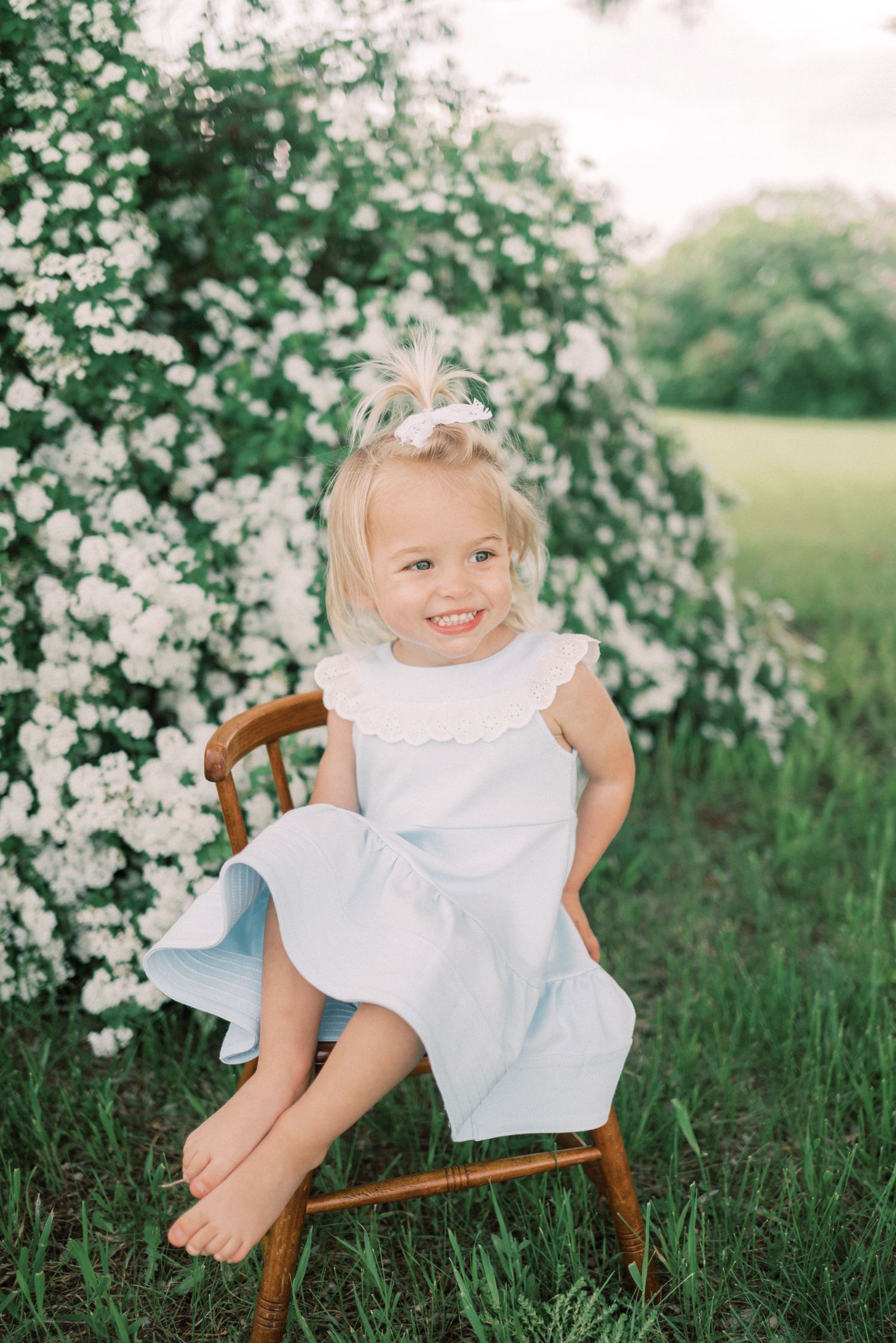 Child in white dress sits on wooden chair against flowering bush backdrop.