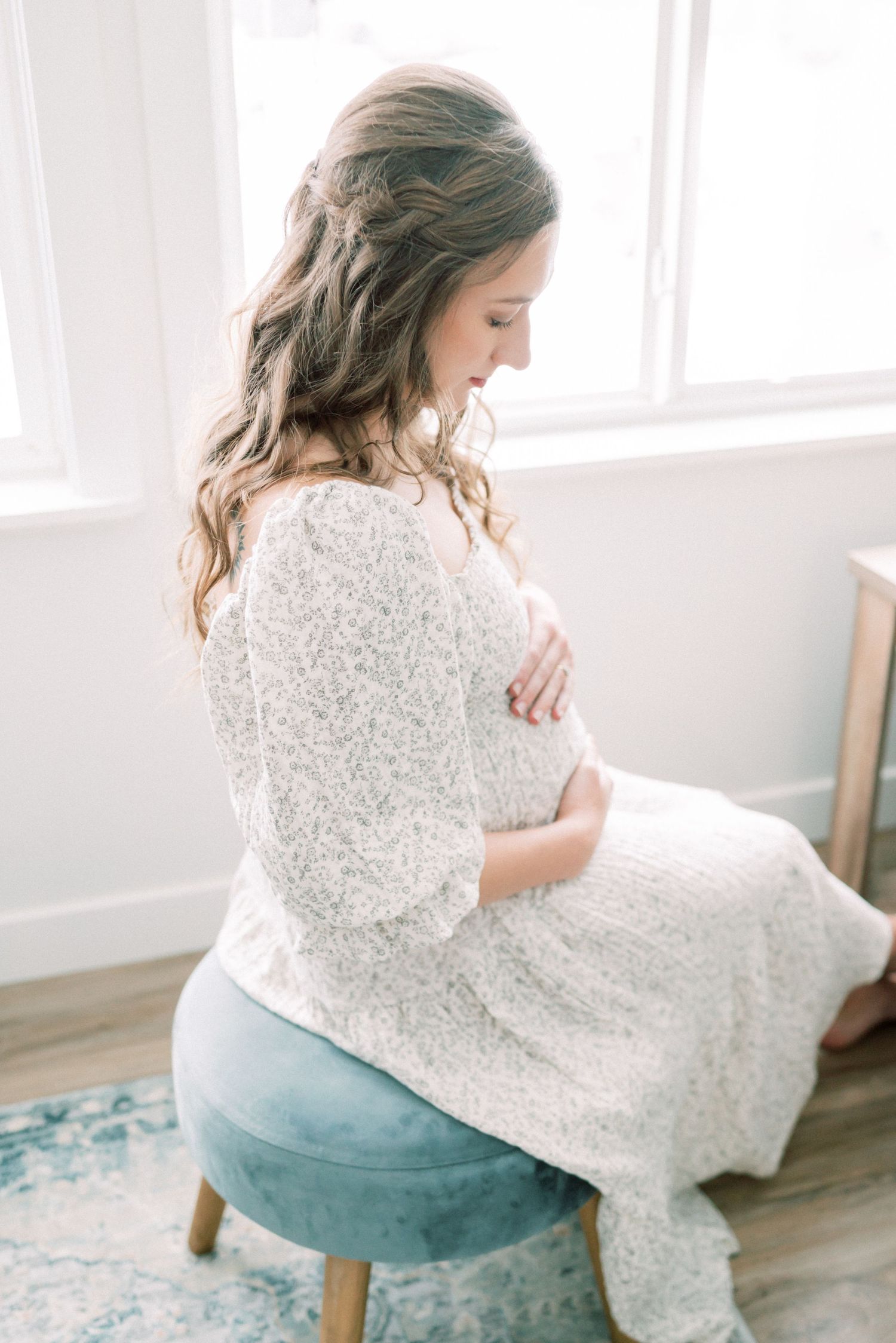 Expecting mother in white lace dress sits on teal chair by window, touching her belly.