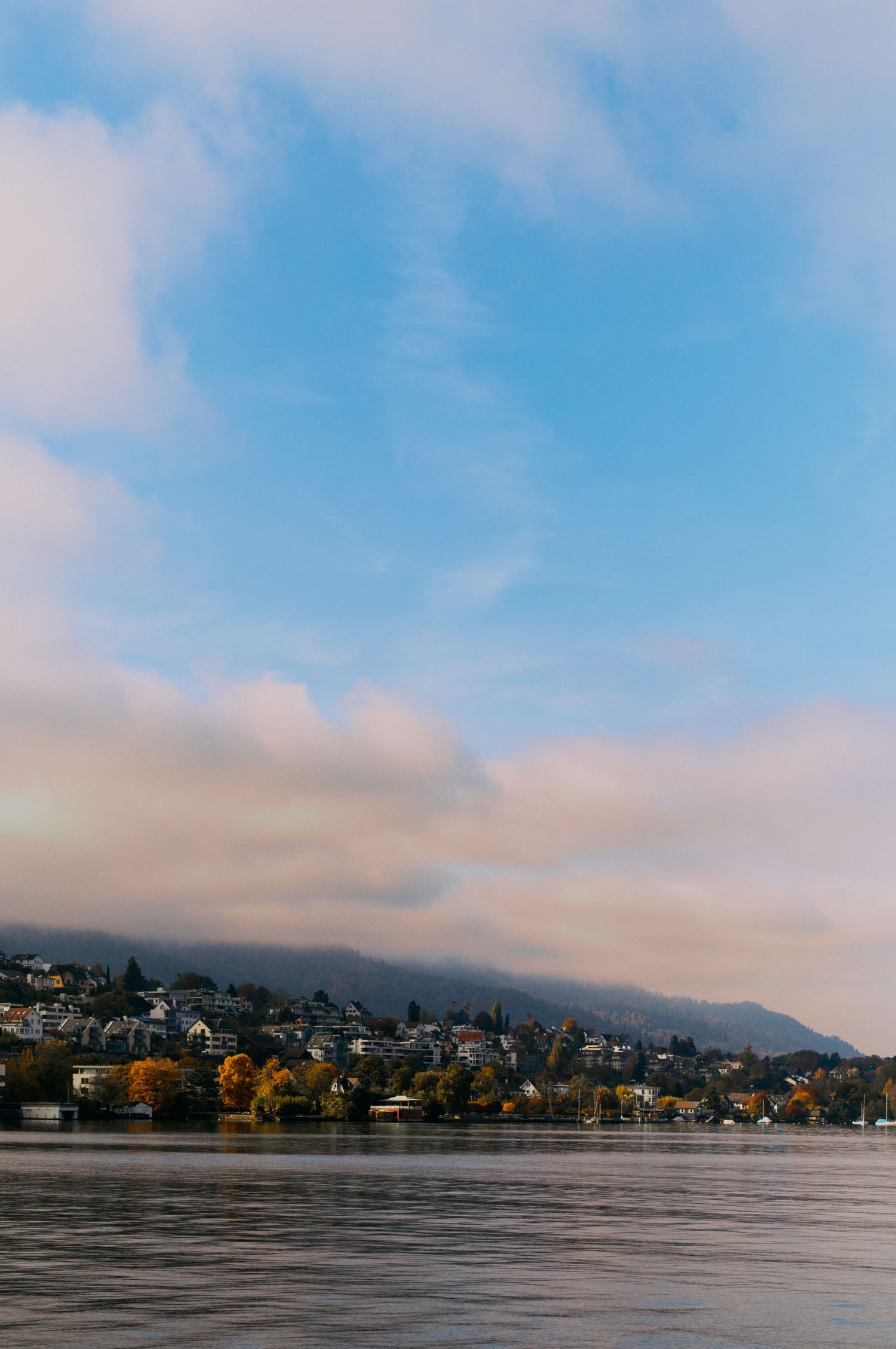 A scenic lakeside town nestled against mountains with autumn trees and moody clouds reflecting on calm waters.