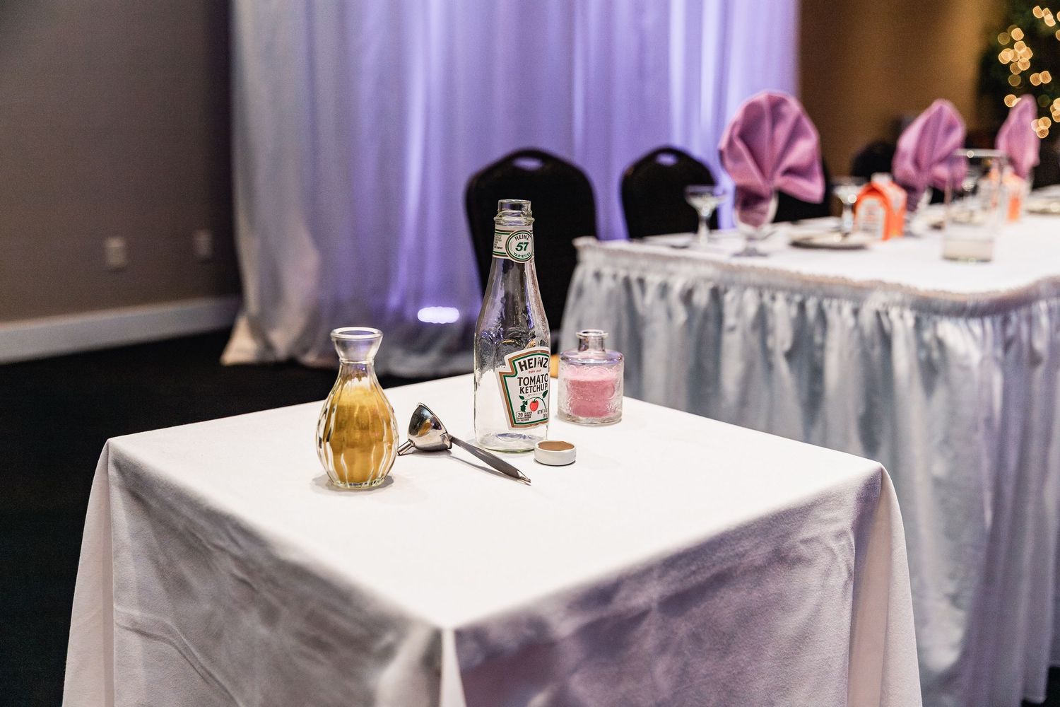 Wedding reception table setting with drinks and white tablecloth, pink napkins visible in background.