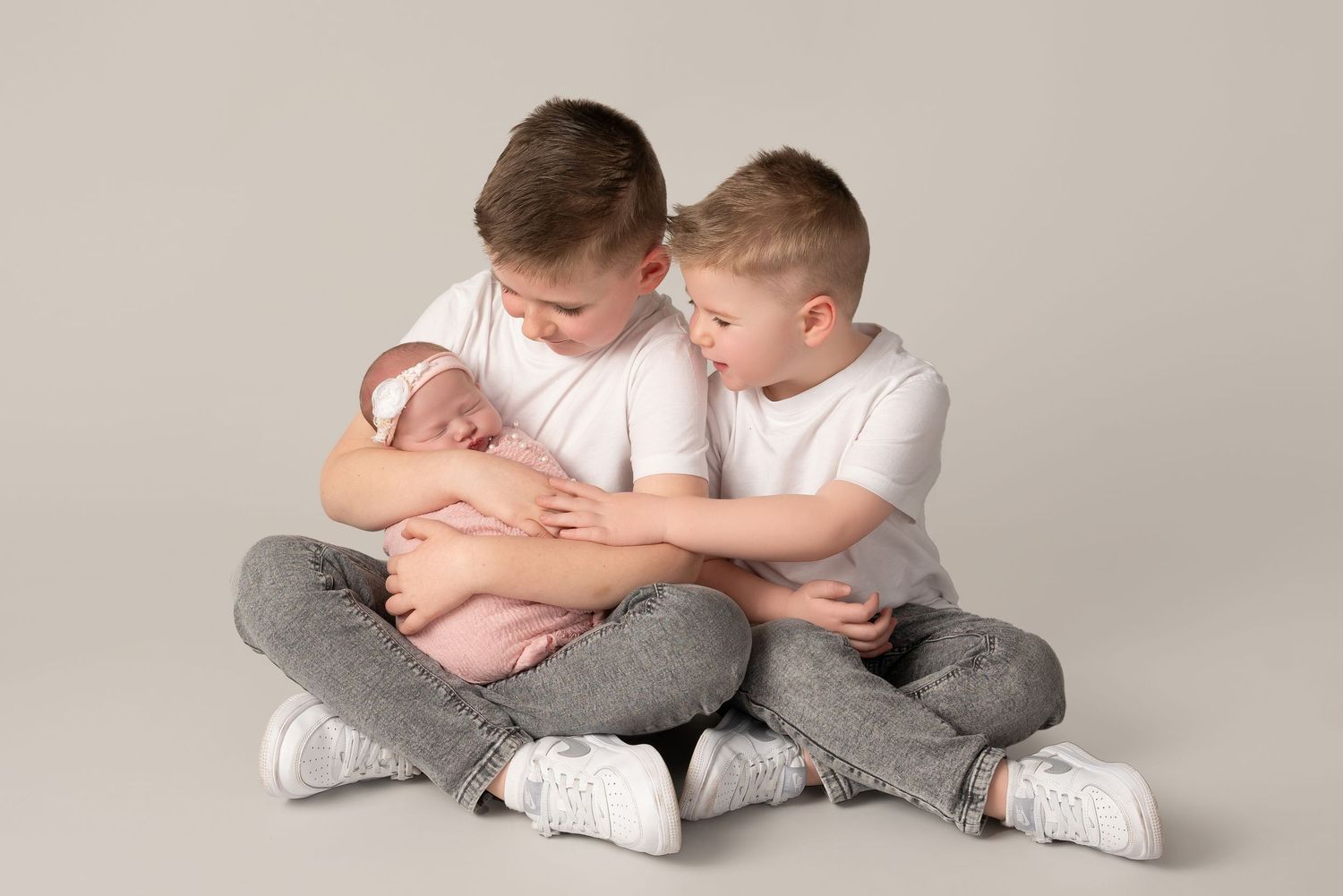 Two siblings in white shirts and gray pants sitting cross-legged while holding a newborn baby on studio backdrop.