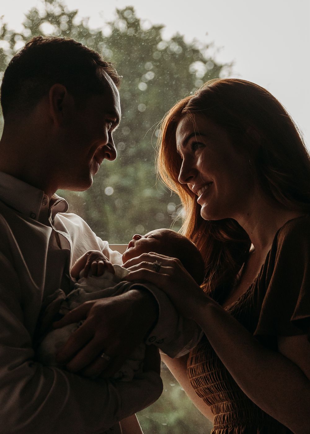 A couple shares a tender moment while bonding with their newborn baby in soft, backlit natural lighting.