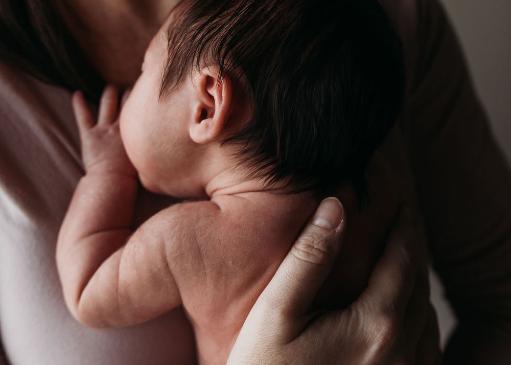 Adult hands gently hold and cradle a sleeping newborn baby against their shoulder in soft lighting.