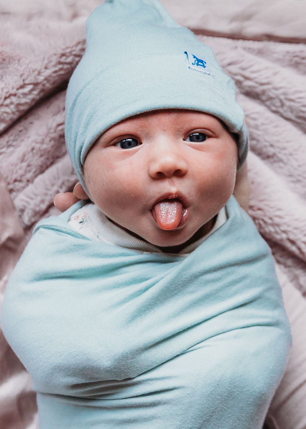 A baby wearing a light blue hat and swaddle blanket sticks out their tongue playfully while lying on soft fabric.
