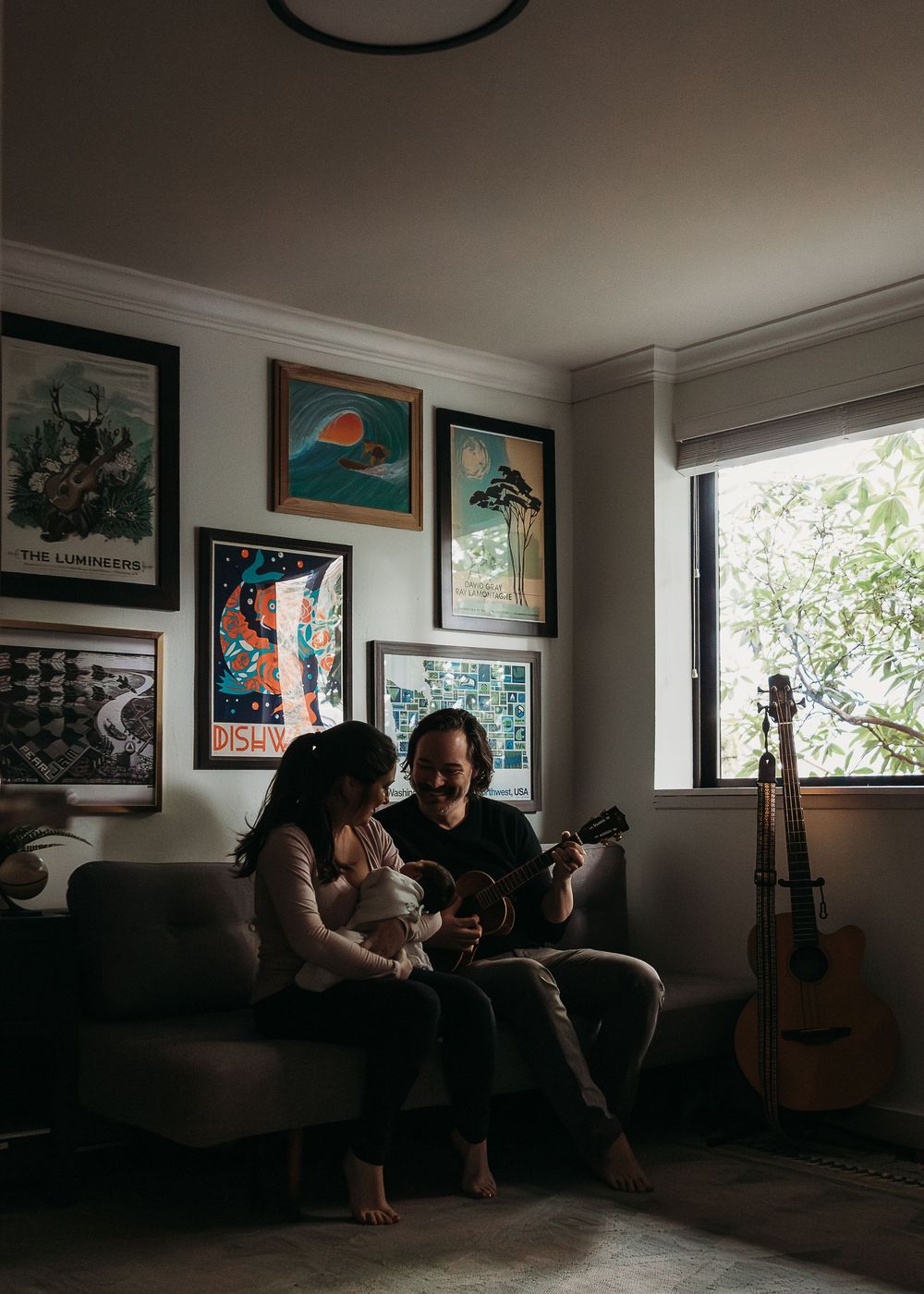 Two people sit on a couch in a cozy room decorated with art prints while playing acoustic guitar near a window.