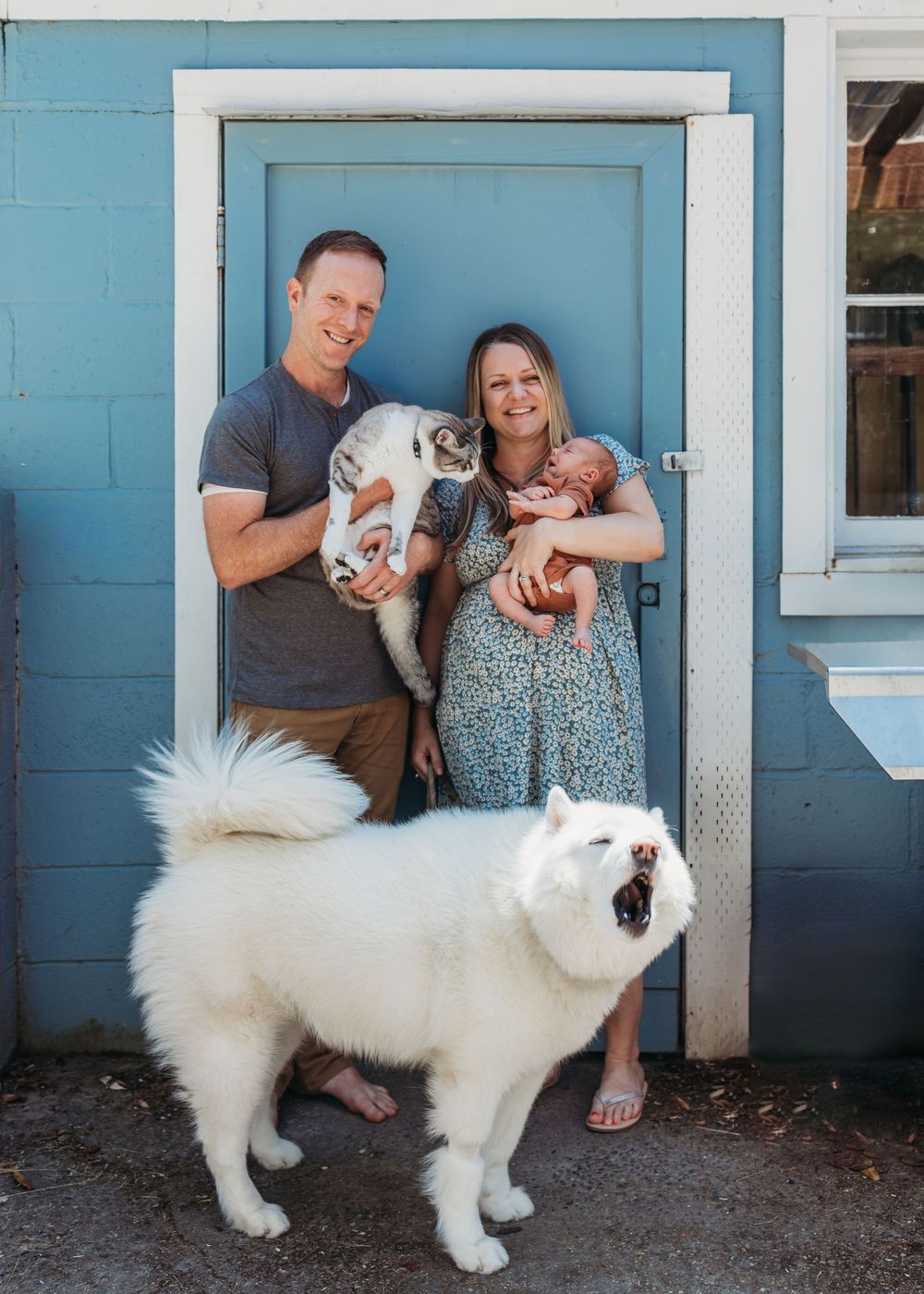 A couple stands by a blue door with two cats while a fluffy white dog yawns in the foreground.