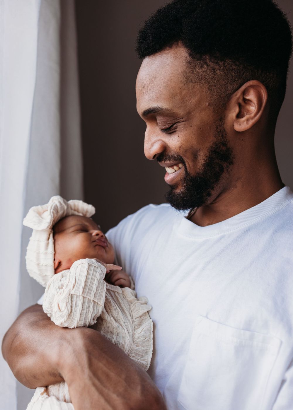 A loving father in a white t-shirt smiles while holding his sleeping newborn baby swaddled in white fabric.
