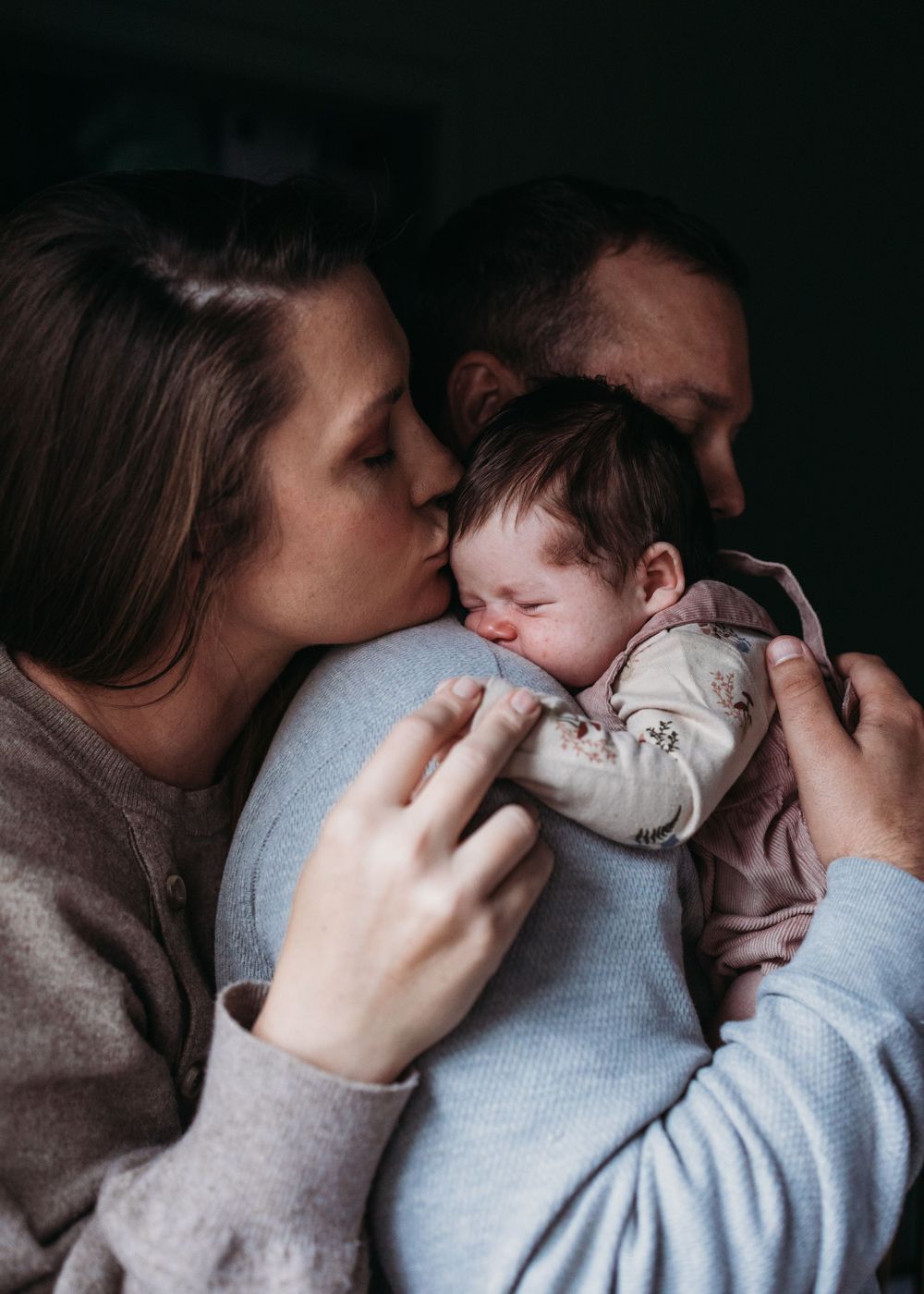 A tender family moment as parents cuddle and kiss their sleepy baby in warm, intimate lighting.