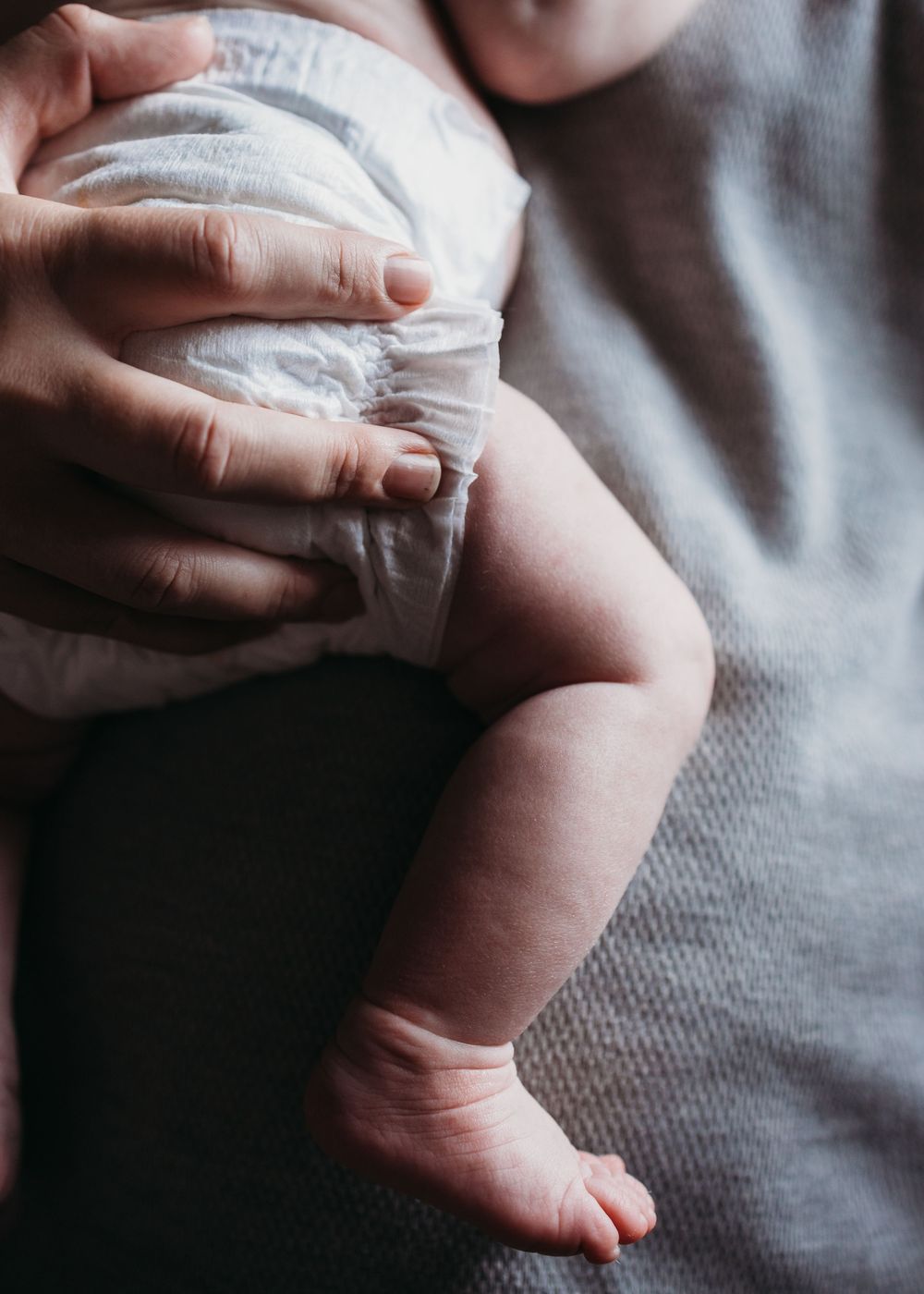 Closeup of tiny baby foot and diaper being held by gentle hands on soft gray fabric background.