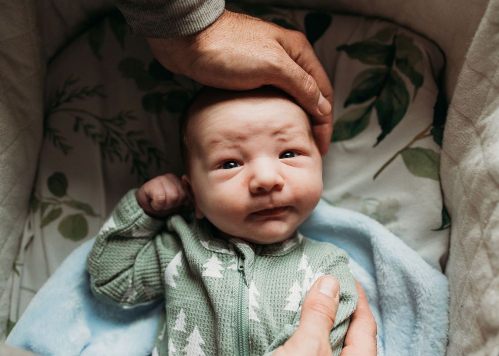 A baby in a mint green cardigan and blue blanket smiles while a hand gently rests on their head.