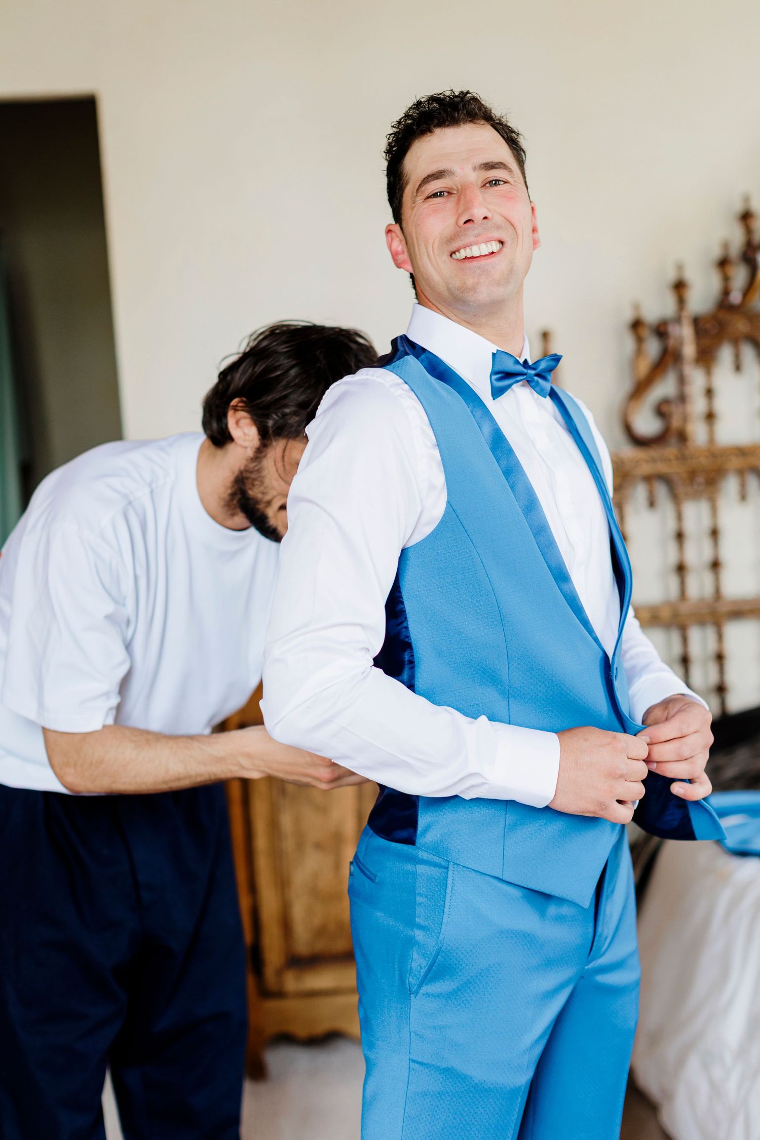 A cheerful person in a powder blue vest and bowtie gets help with formal attire.