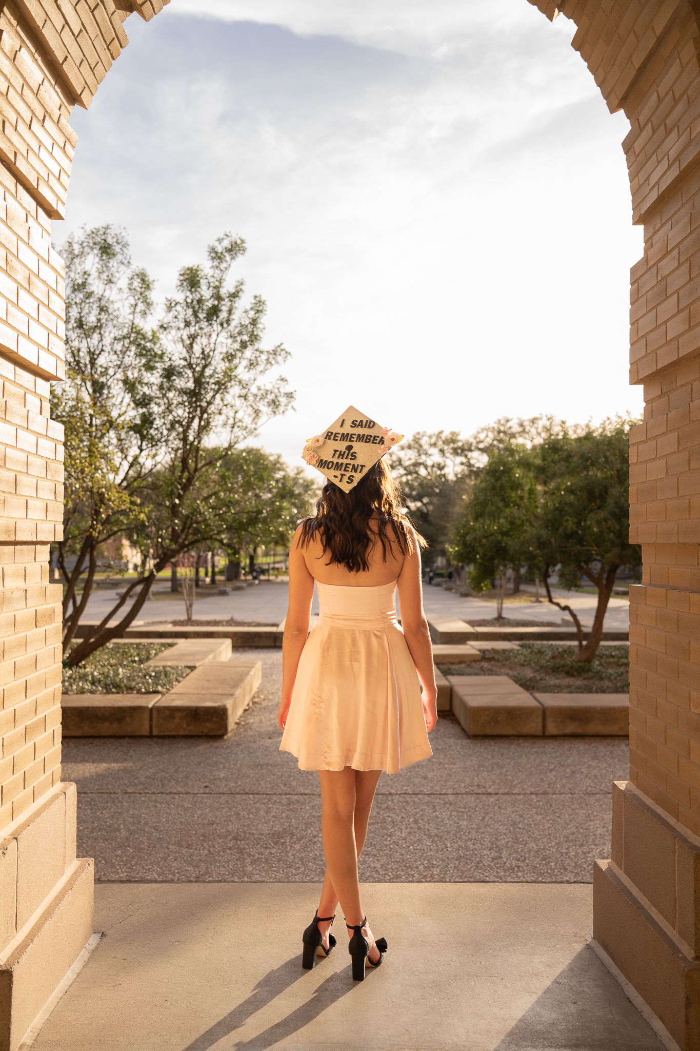 Graduate in yellow sundress and cap stands beneath stone archway on college campus at sunset.