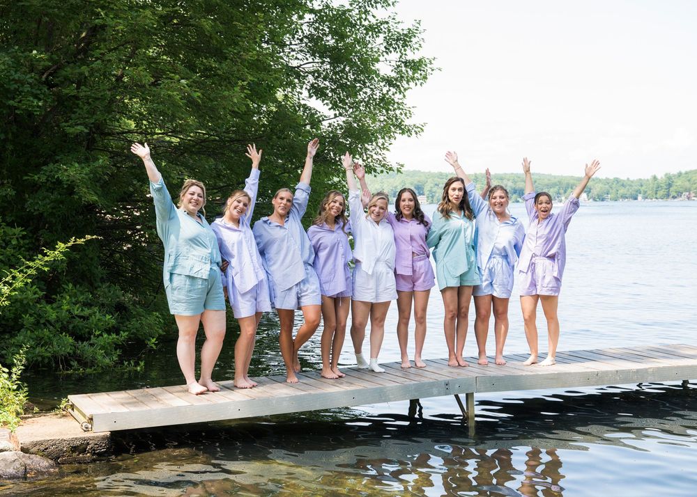 Group of bridesmaids in pastel dresses celebrating on a dock by a lake.