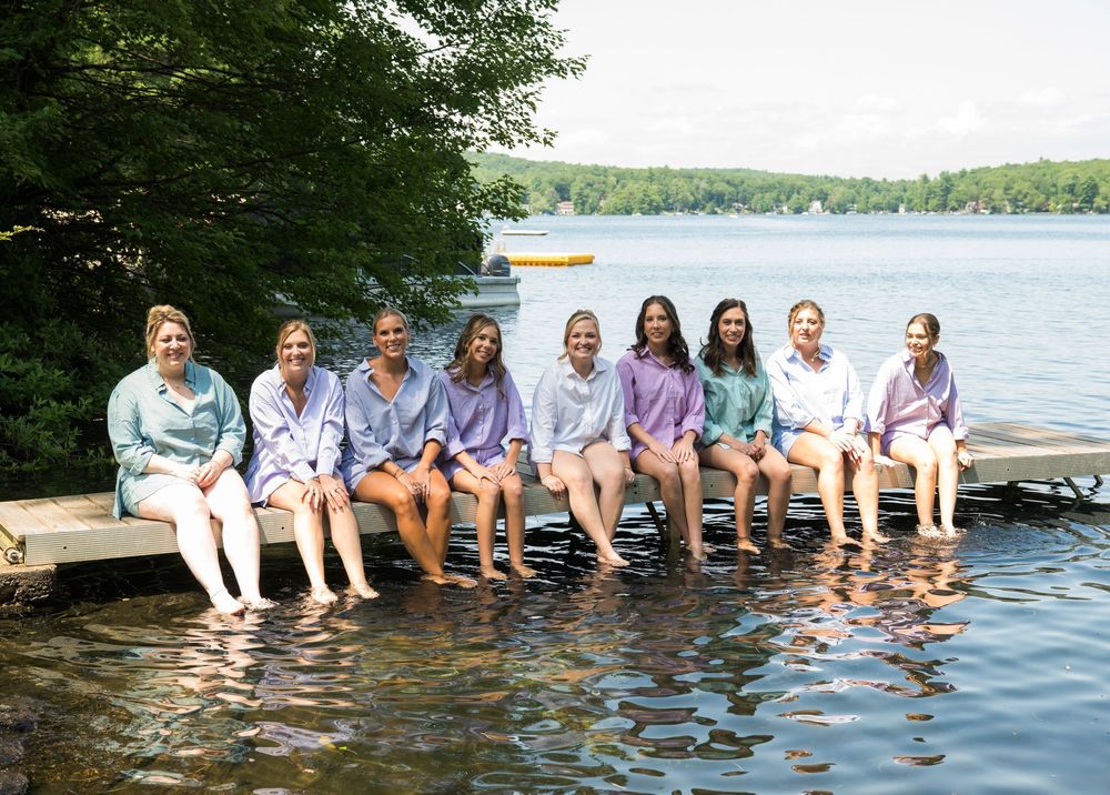 A group of people in color-coordinated blue and purple outfits sitting on a dock with their feet in a peaceful lake.
