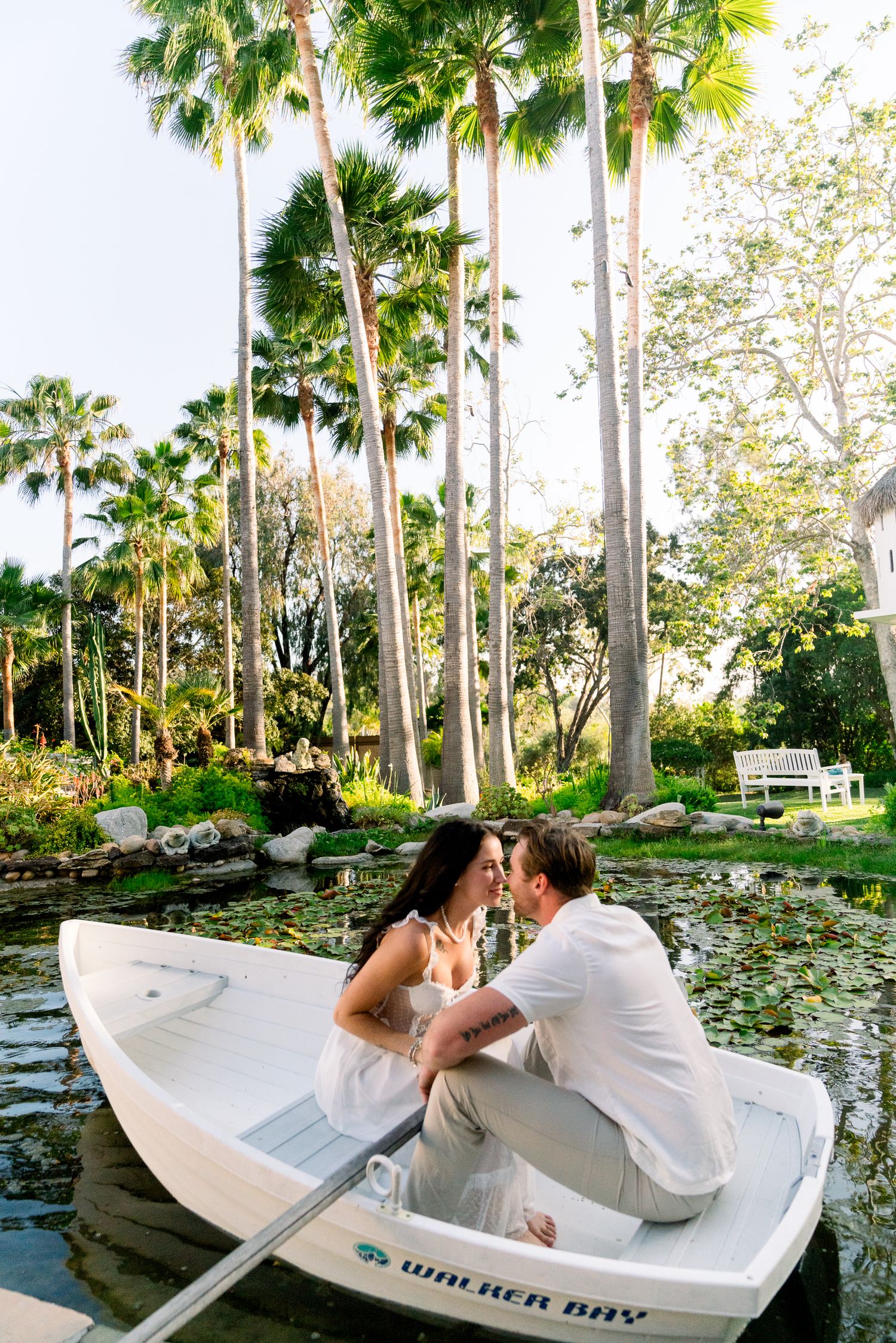 Two people share an intimate moment while sitting in a white rowboat on a pond surrounded by tropical palm trees.
