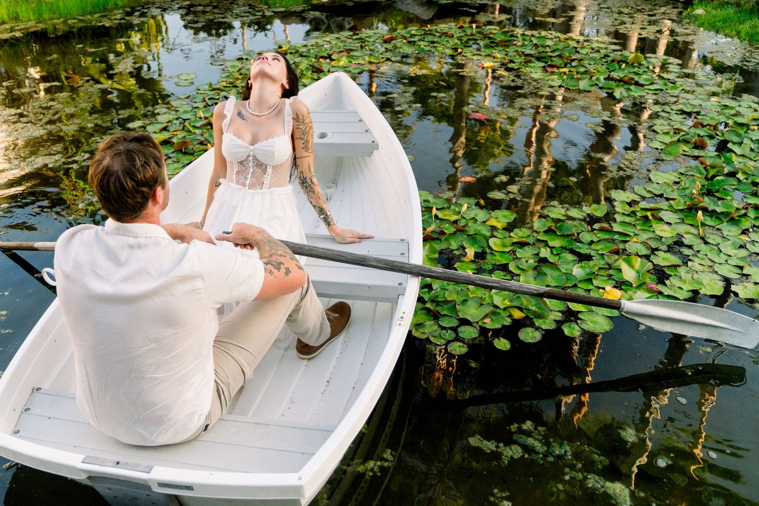 A romantic scene unfolds as a couple sits in a white boat surrounded by lily pads on a tranquil pond.
