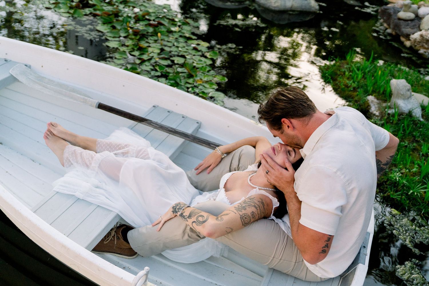A couple shares a romantic moment while lying in a white rowboat surrounded by greenery and water.