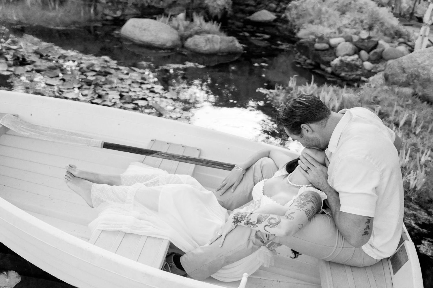 A couple shares an intimate moment in black and white while sitting in a small rowboat on a peaceful lake.