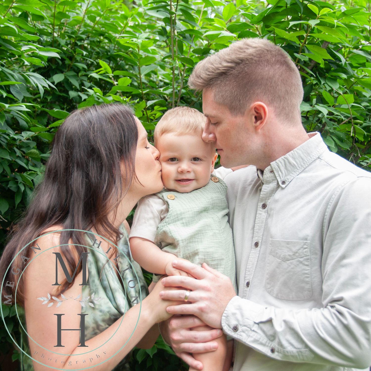 Image of a family of three - mom and dad kissing a little boy outdoors with green foliage background.