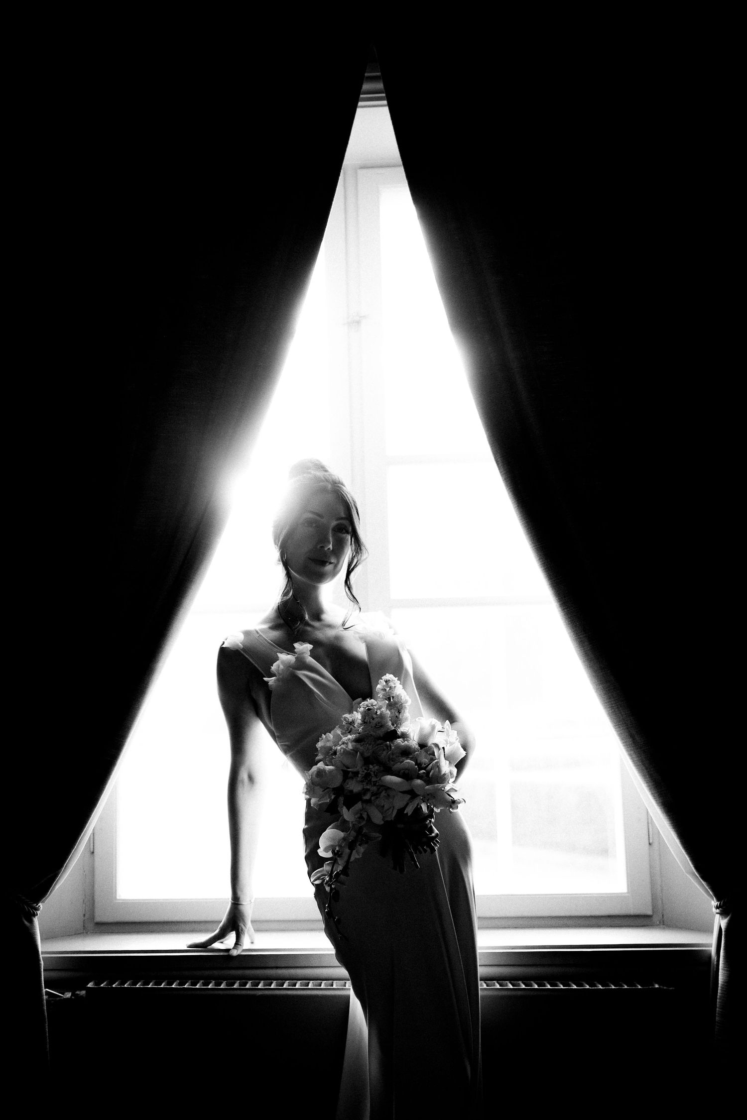 Black and white silhouette of an elegant figure holding a bouquet against a bright triangular window in dramatic lighting.