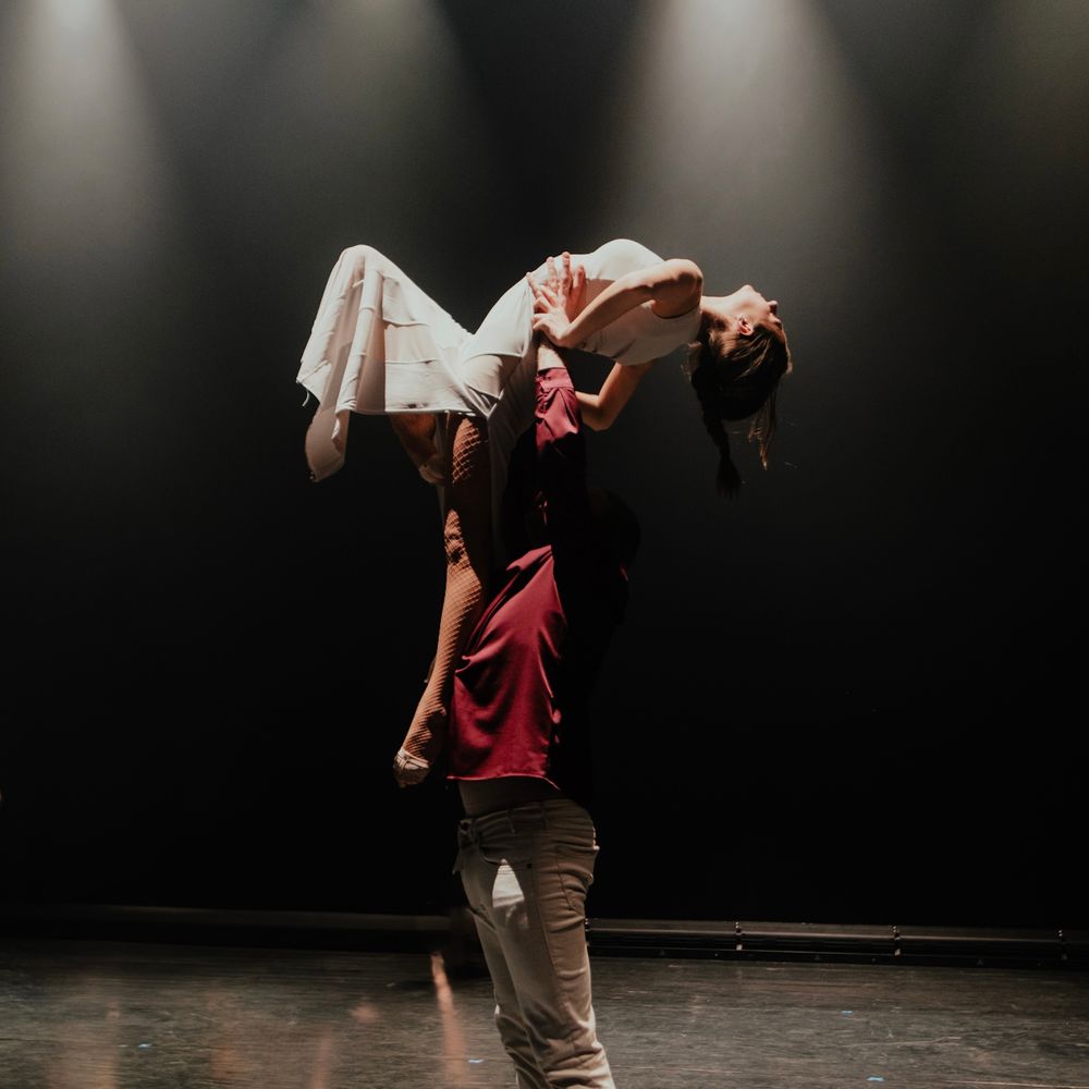 Two dancers performing a dramatic lift on a dark stage with one elevated horizontally in an artistic pose.