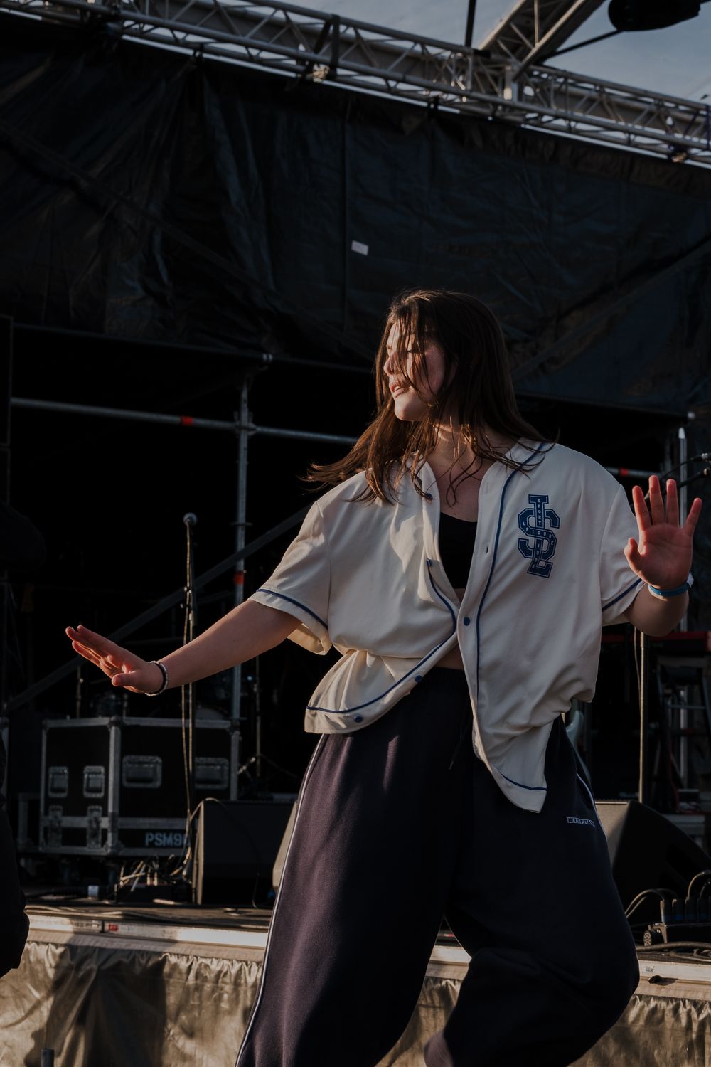 Performer in a white baseball jersey energetically dancing on an outdoor festival stage during a sunny day.