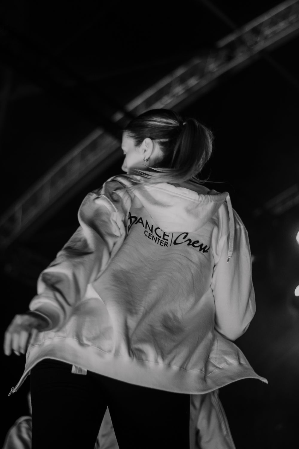 Black and white photo of a dancer wearing a white dance crew jacket shown from behind while performing.