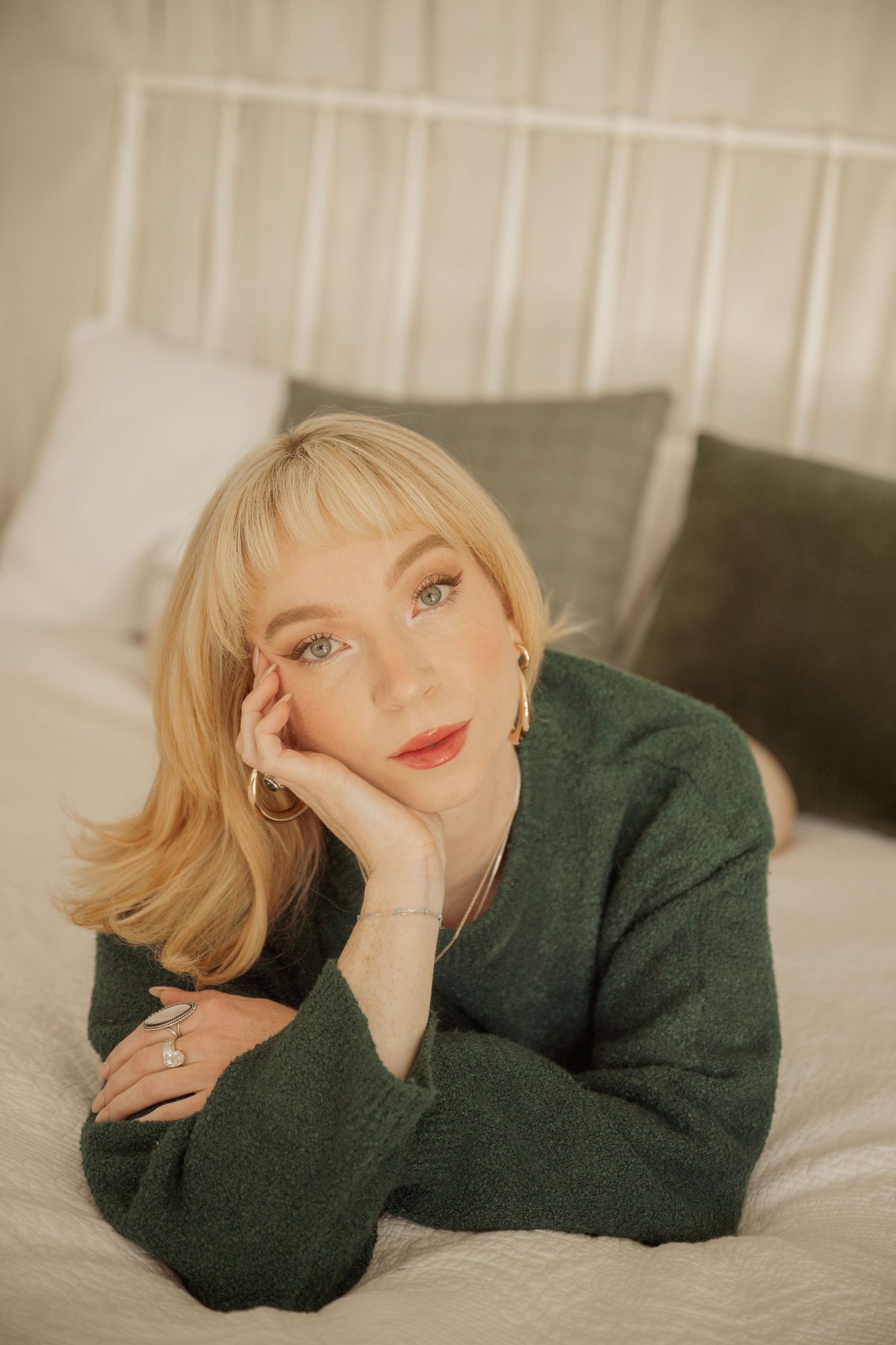 Someone in a green sweater poses on a bed with pillows in a warm, cozy indoor setting.