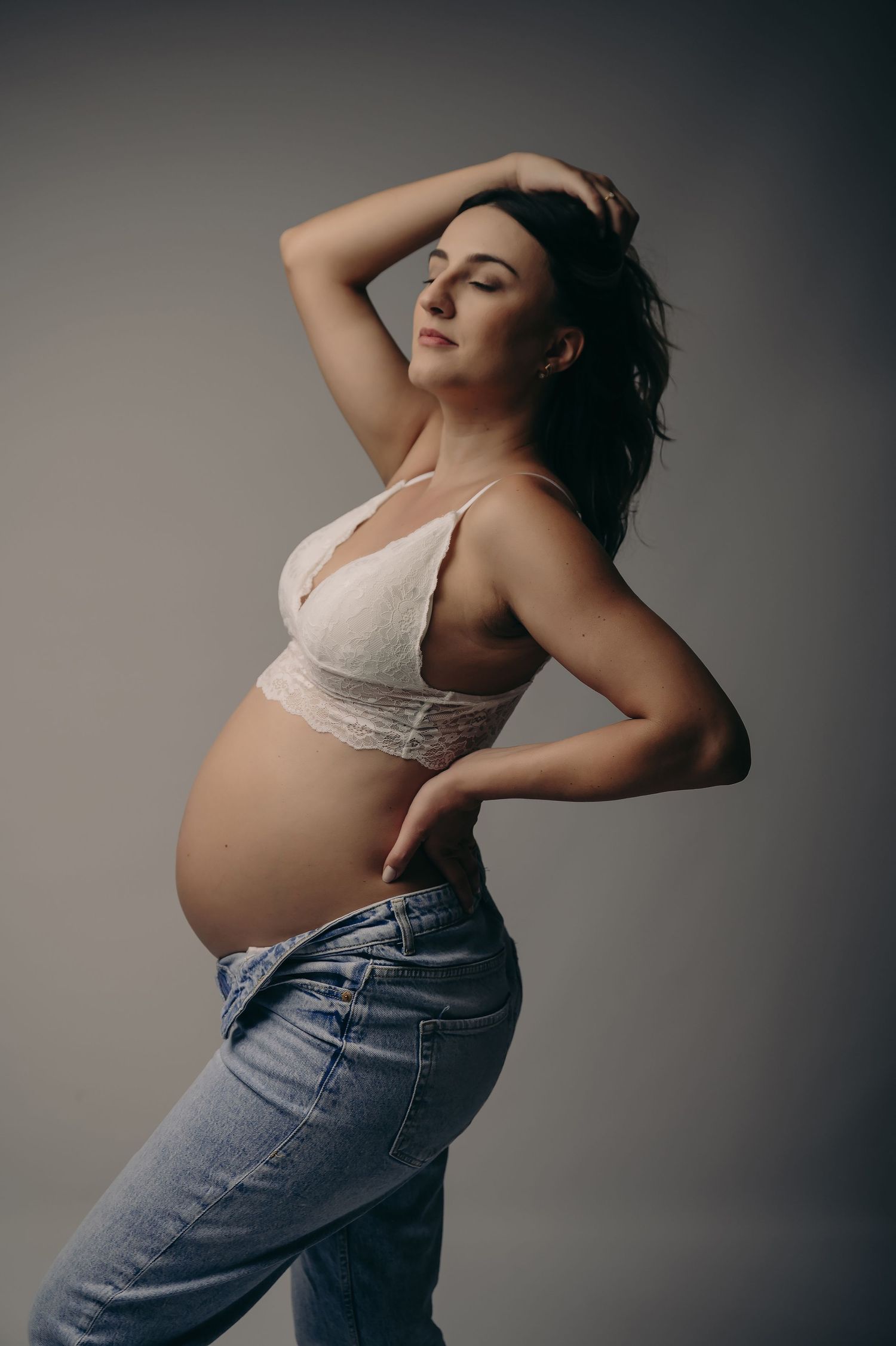 Artistic maternity portrait of an expectant mother in lace bralette and jeans posing against a neutral background.