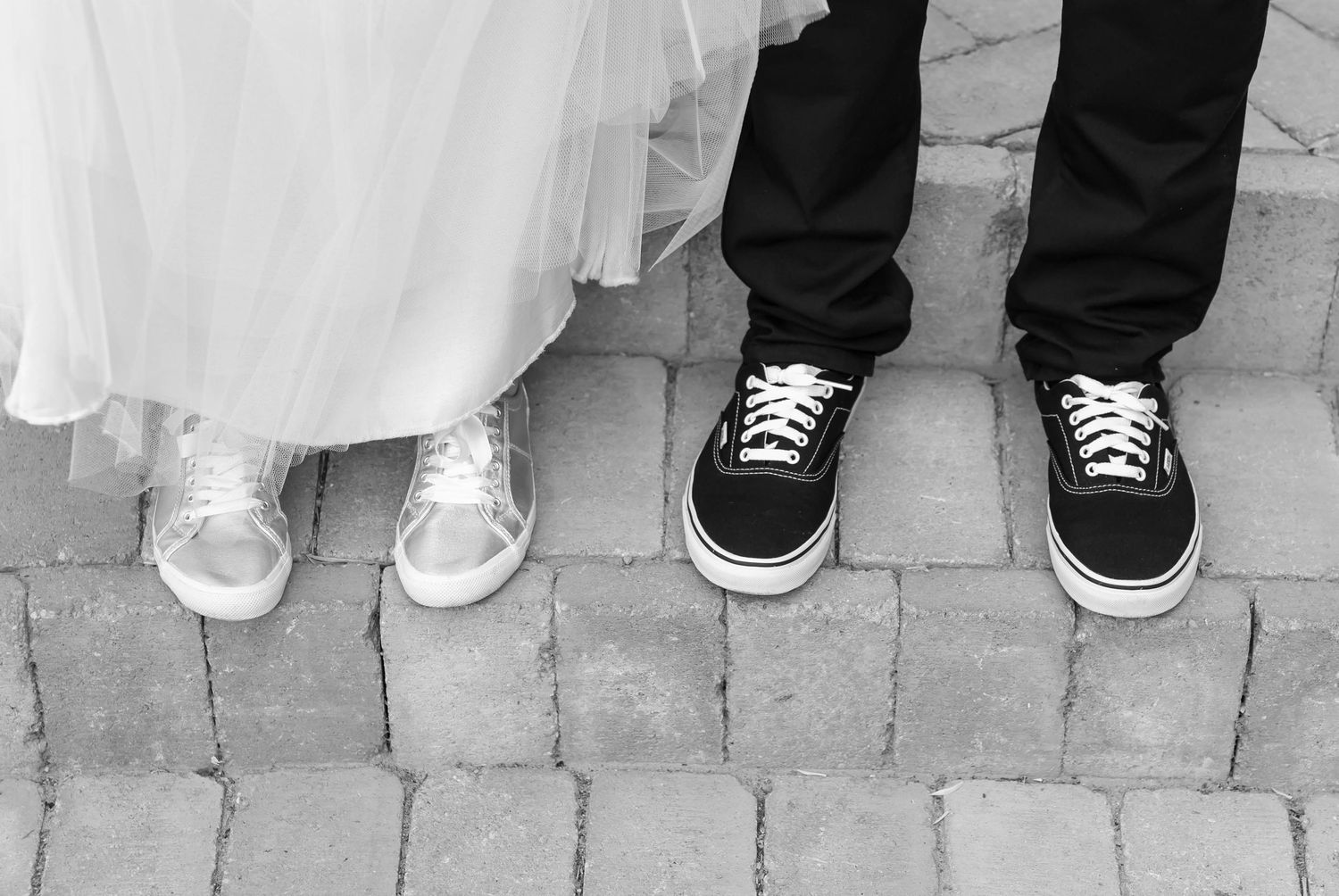 A black and white photo of bride and groom's feet showing sneakers, with the bride in white and groom in black shoes.