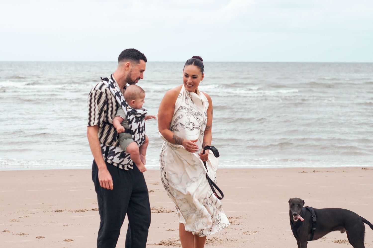 Two beachgoers and their black dog walk along a sandy shore with waves crashing in the background.