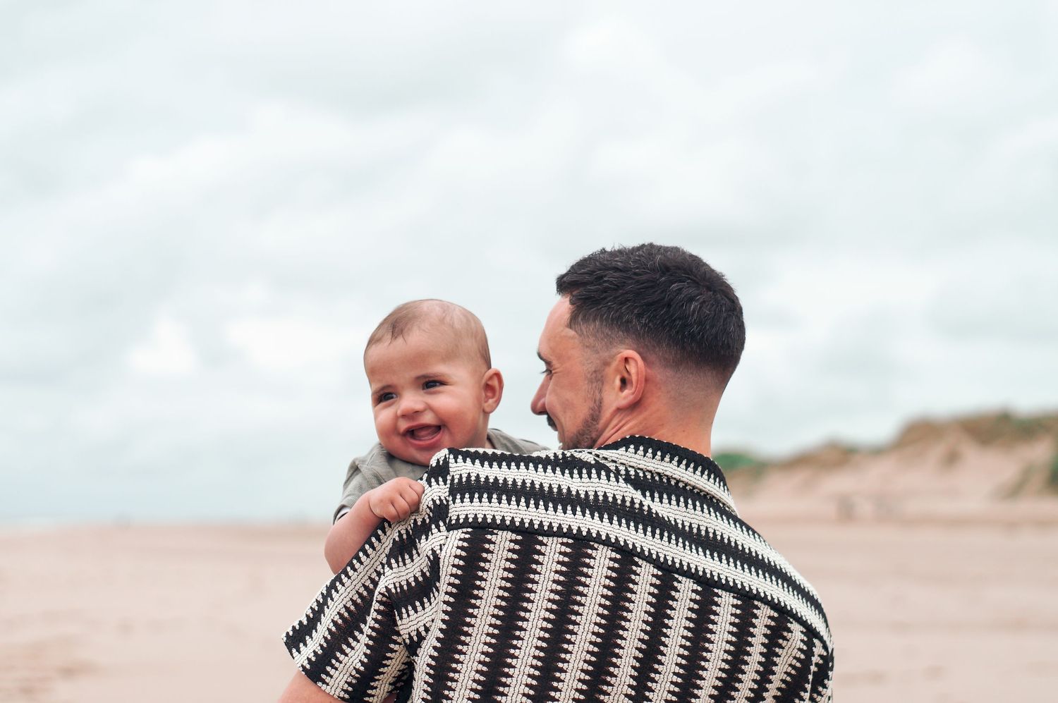 Person wearing a black and white striped sweater holds a smiling baby on a sandy beach.
