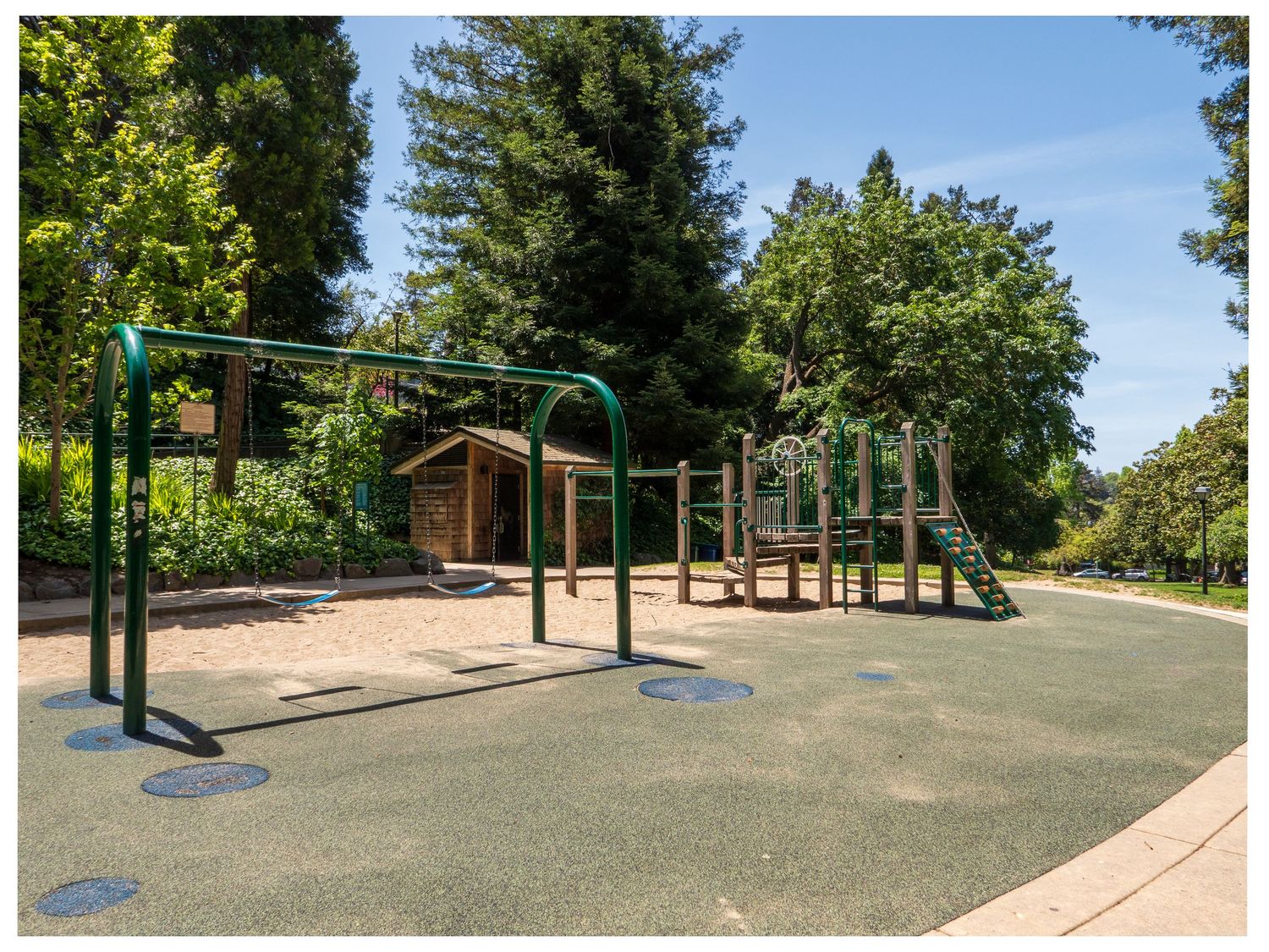 Empty playground equipment with swing sets and climbing structure on rubber surface in a sunny park setting surrounded by trees.