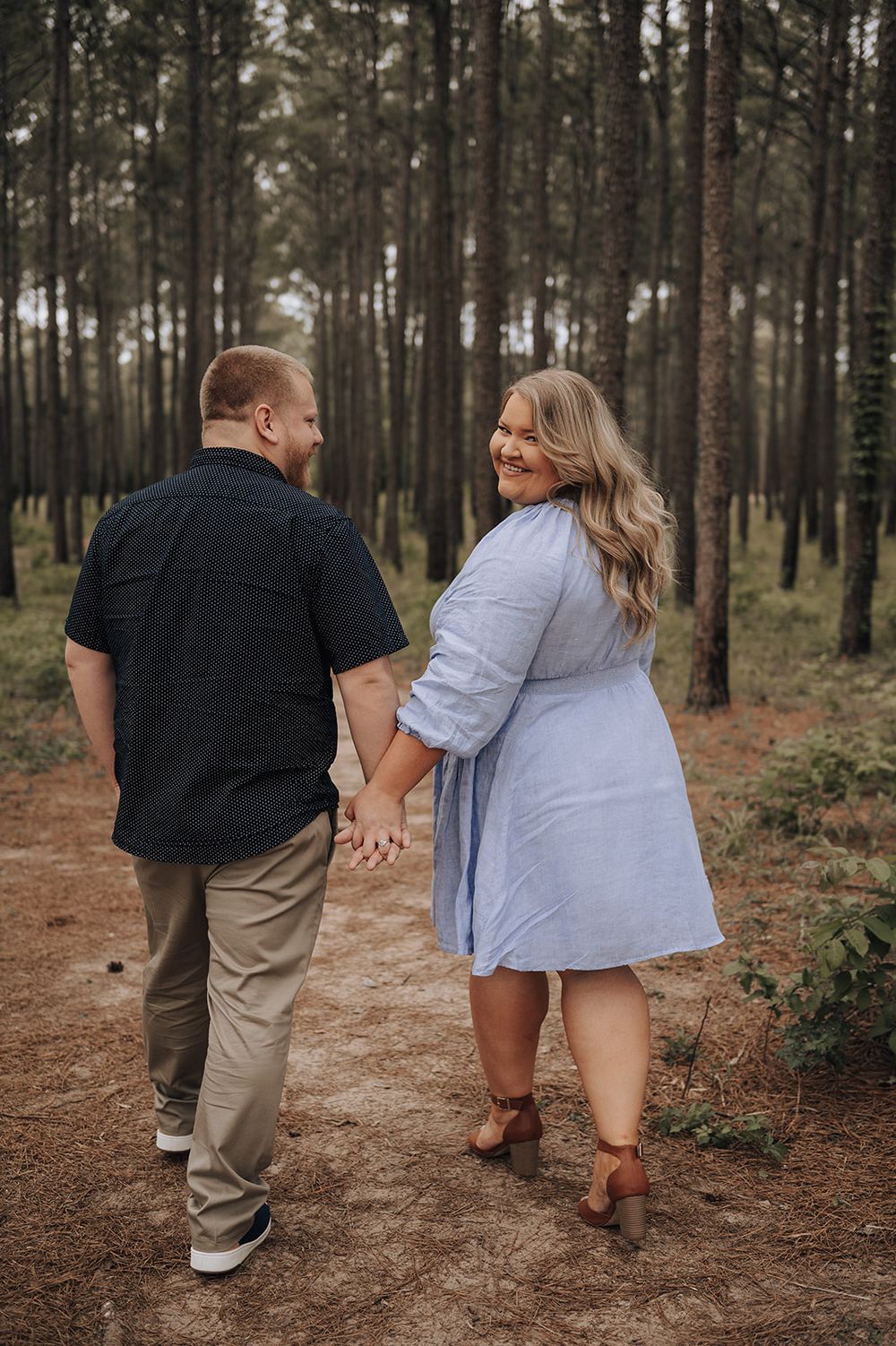 A couple holding hands walking down a forest path in casual clothing surrounded by tall pine trees on a misty day.