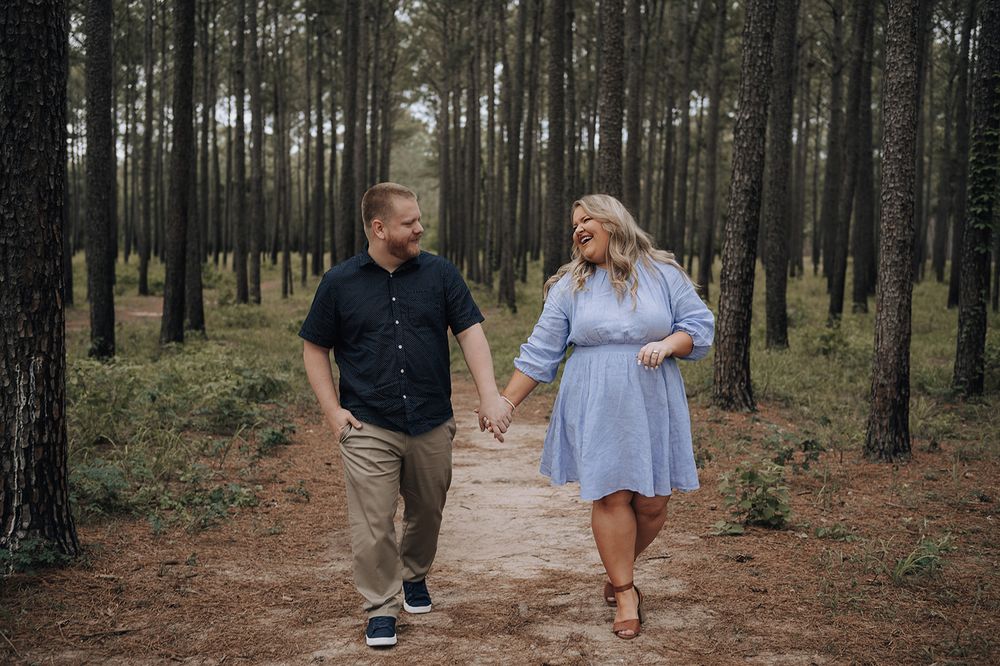 A couple walks hand in hand on a dirt path through a serene pine forest while sharing a joyful moment together.