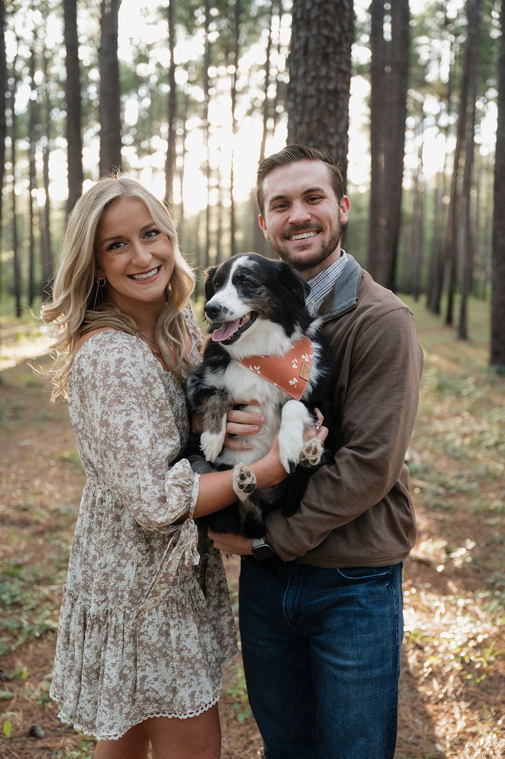 A couple holding their black and white dog poses together in a sunlit forest wearing casual fall attire.