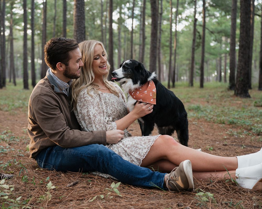 Couple sits together on forest floor with their black and white dog in a serene pine forest setting.