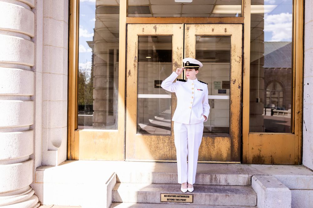 Naval officer in dress whites renders a salute on stone steps of a columned building entrance.